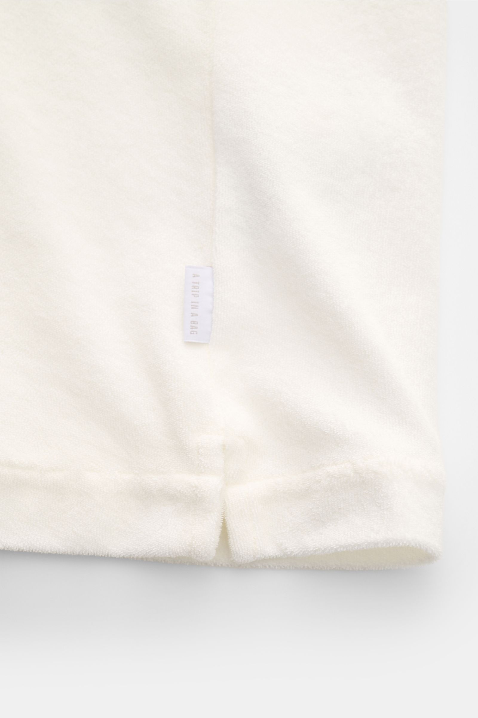 Close-up front view of 04651/ A trip in a bag Frottee-Kurzarmhemd 'Terry Cuba' offwhite hem with small side slit and fabric label.

Description: Urlaubsgefühl zum Anziehen: Das Kurzarmhemd 'Terry Cuba' von 04651/ A TRIP IN A BAG aus angenehmem Baumwoll