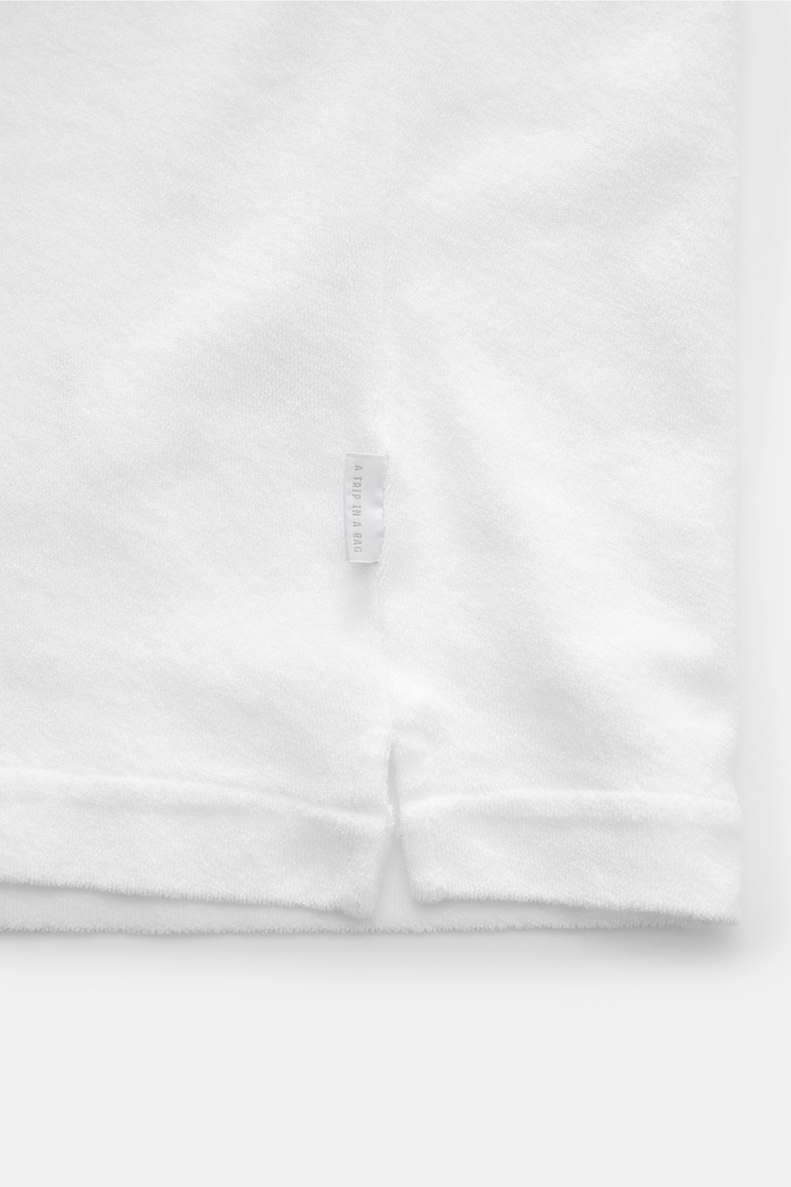 White terry cotton fabric close-up showing texture, small side seam label, and hem slit from a top-front perspective of the 04651/ A trip in a bag Frottee-Kurzarmhemd 'Terry Cuba' weiß. Urlaubsgefühl zum Anziehen: Das Kurzarmhemd 'Terry Cuba' von 04651/ A
