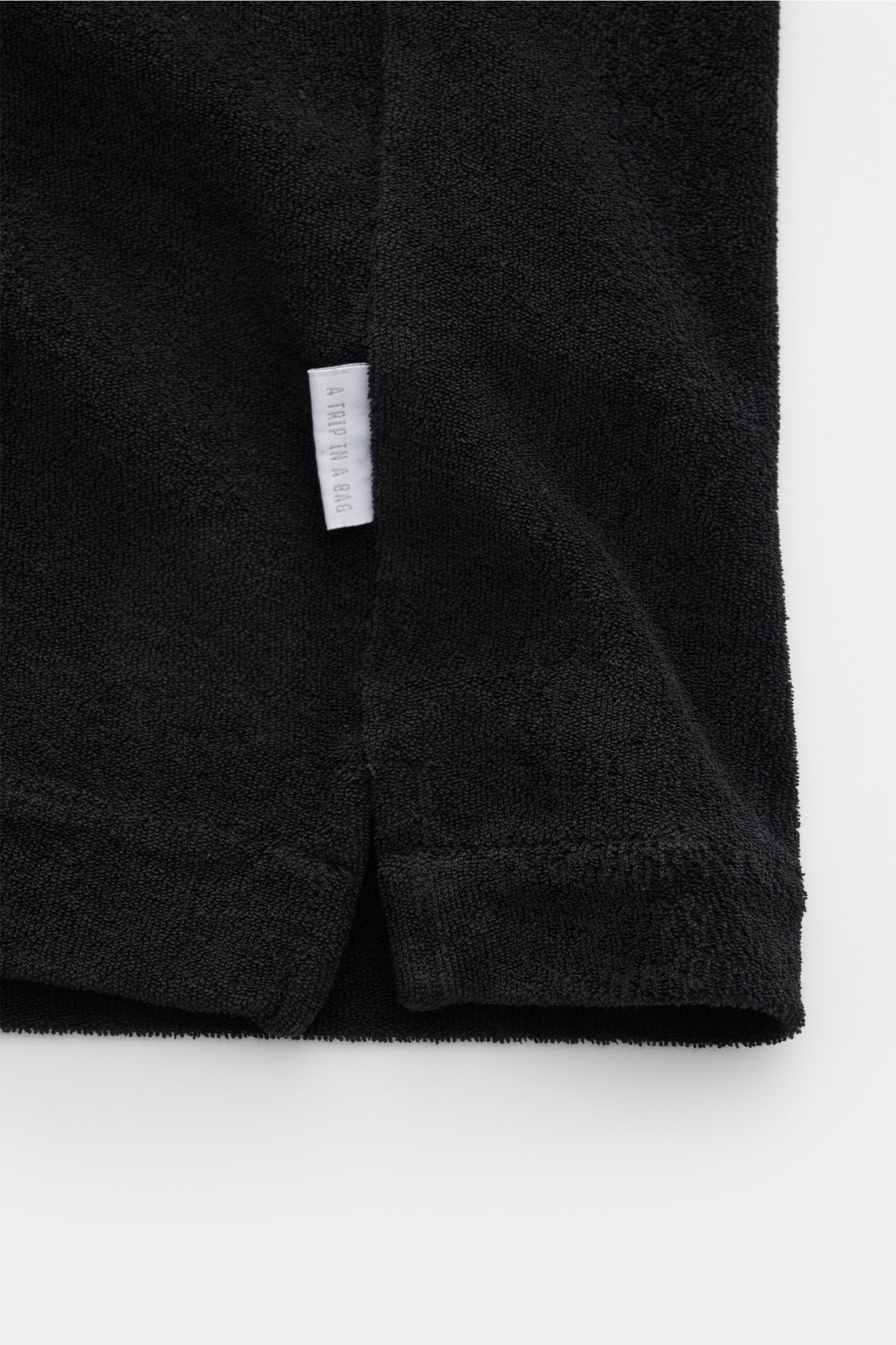 Schwarzer Saum mit kurzen Schlitzen und kleinem Weblabel, Detailfoto aus Nahaufnahme des 04651/ A trip in a bag Frottee-Kurzarmhemd 'Terry Cuba' schwarz aus Baumwoll-Frottee.