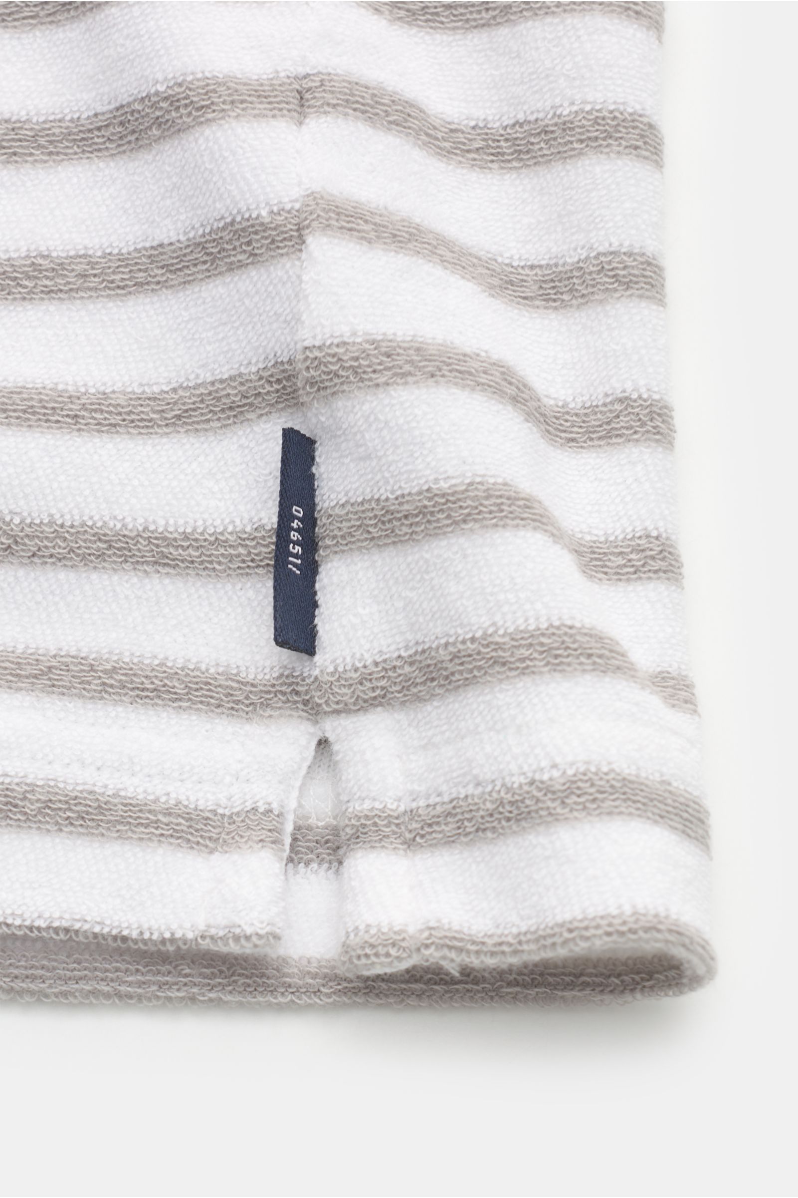 Nahaufnahme des Saum mit kurzem Schlitz und kleinem Weblabel des 04651/ A trip in a bag Frottee-Poloshirt 'Terry Stripe Polo' grau/weiß gestreift aus weichem Baumwoll-Frottee.