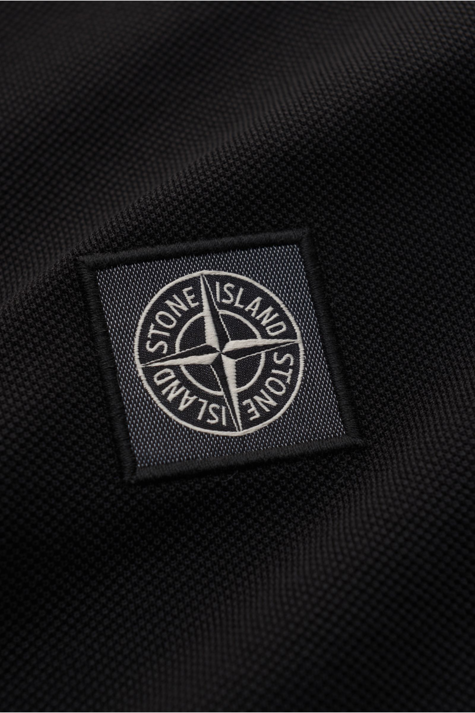 Close-up of the Stone Island Poloshirt schwarz fabric showing the detailed black cotton piqué texture and the stitched Stone Island logo patch from a front angle. The classic polo shirt is a must-have in the sporty men's wardrobe: made of pure cotton piqu