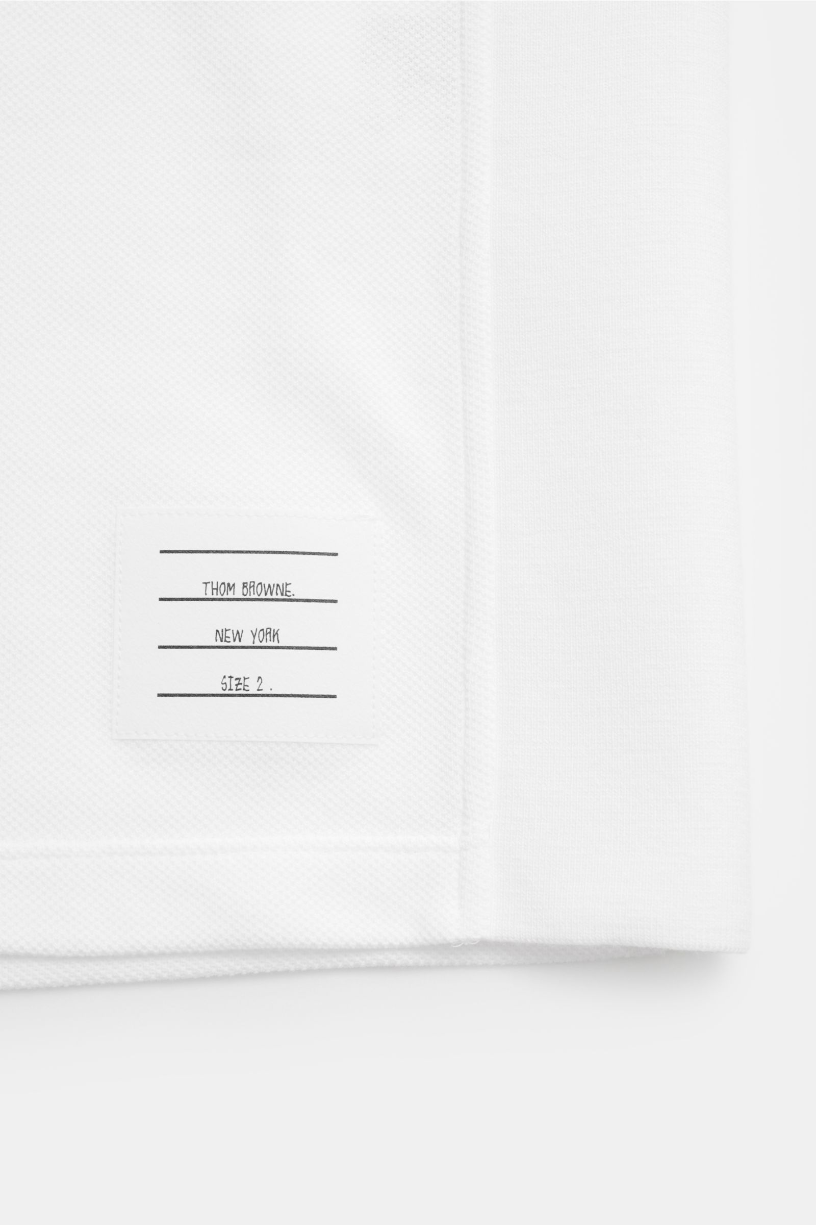 Close-up of the lower side hem of the Thom Browne Polo shirt white, showing the signature label and textured pure cotton fabric from a front angle.

Description to include: 
A modern essential: This polo shirt by THOM BROWNE is luxurious and comfortabl