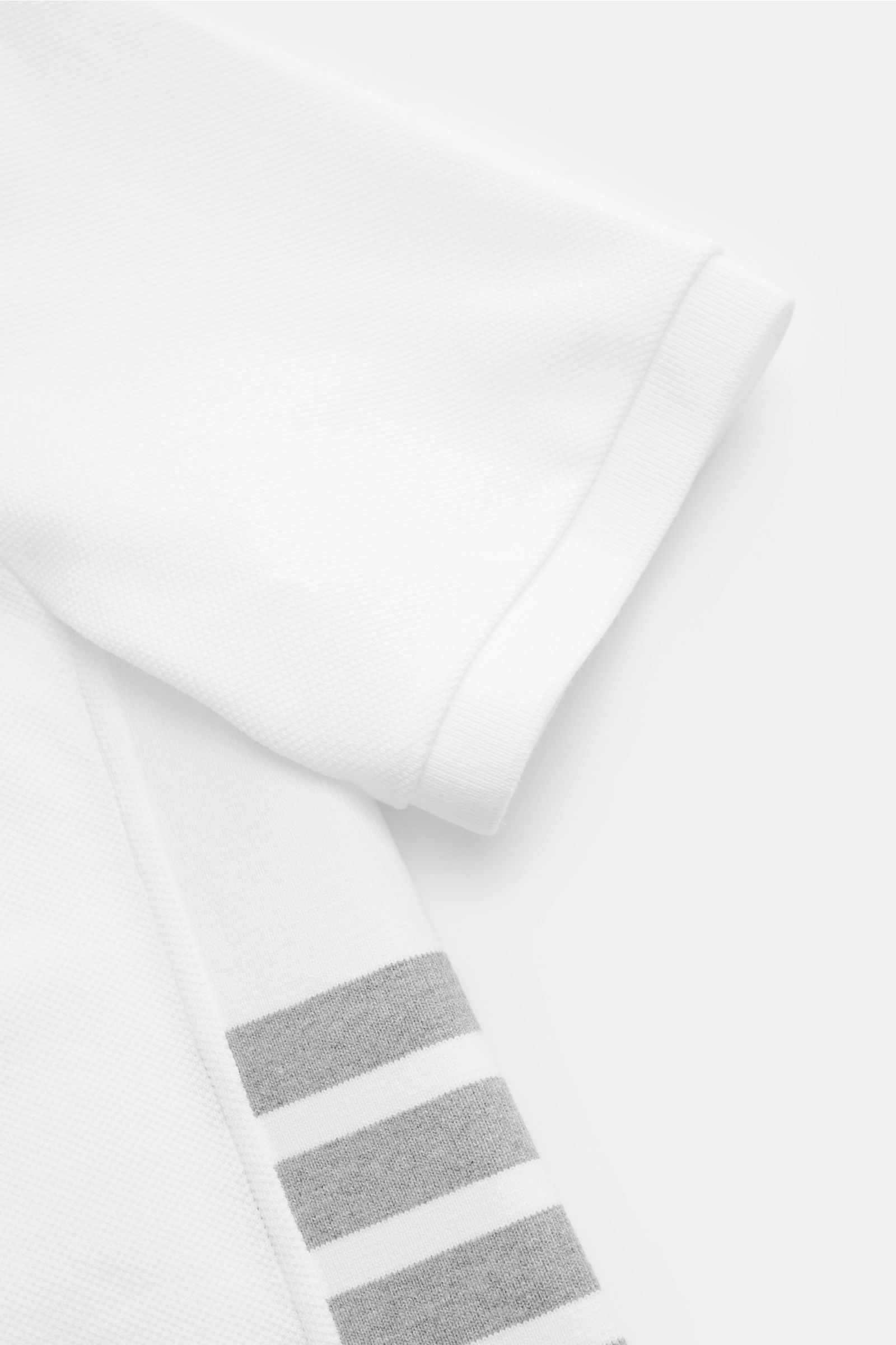 Close-up side view of Thom Browne Polo shirt white sleeve showing pure cotton fabric, slim fit, ribbed cuff, stretchy side inserts, and signature striped logo. A modern essential: This polo shirt by THOM BROWNE is luxurious and comfortable, which makes