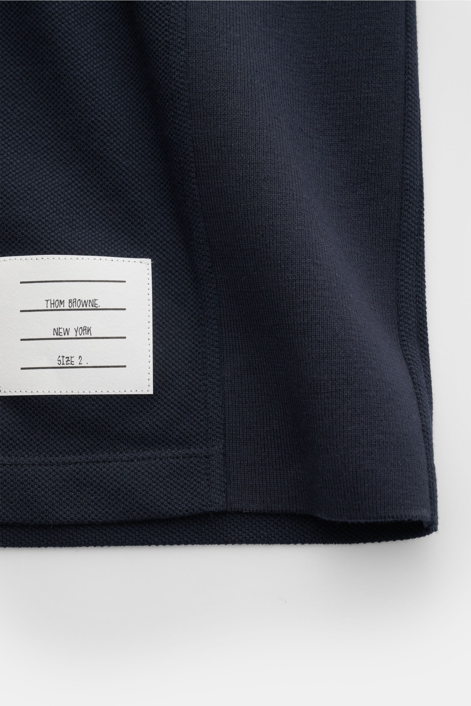 Nahaufnahme der unteren Saumkante des Thom Browne Poloshirts navy aus reiner Baumwolle, mit markentypischem weißen Label, gezeigt von unten vorne.