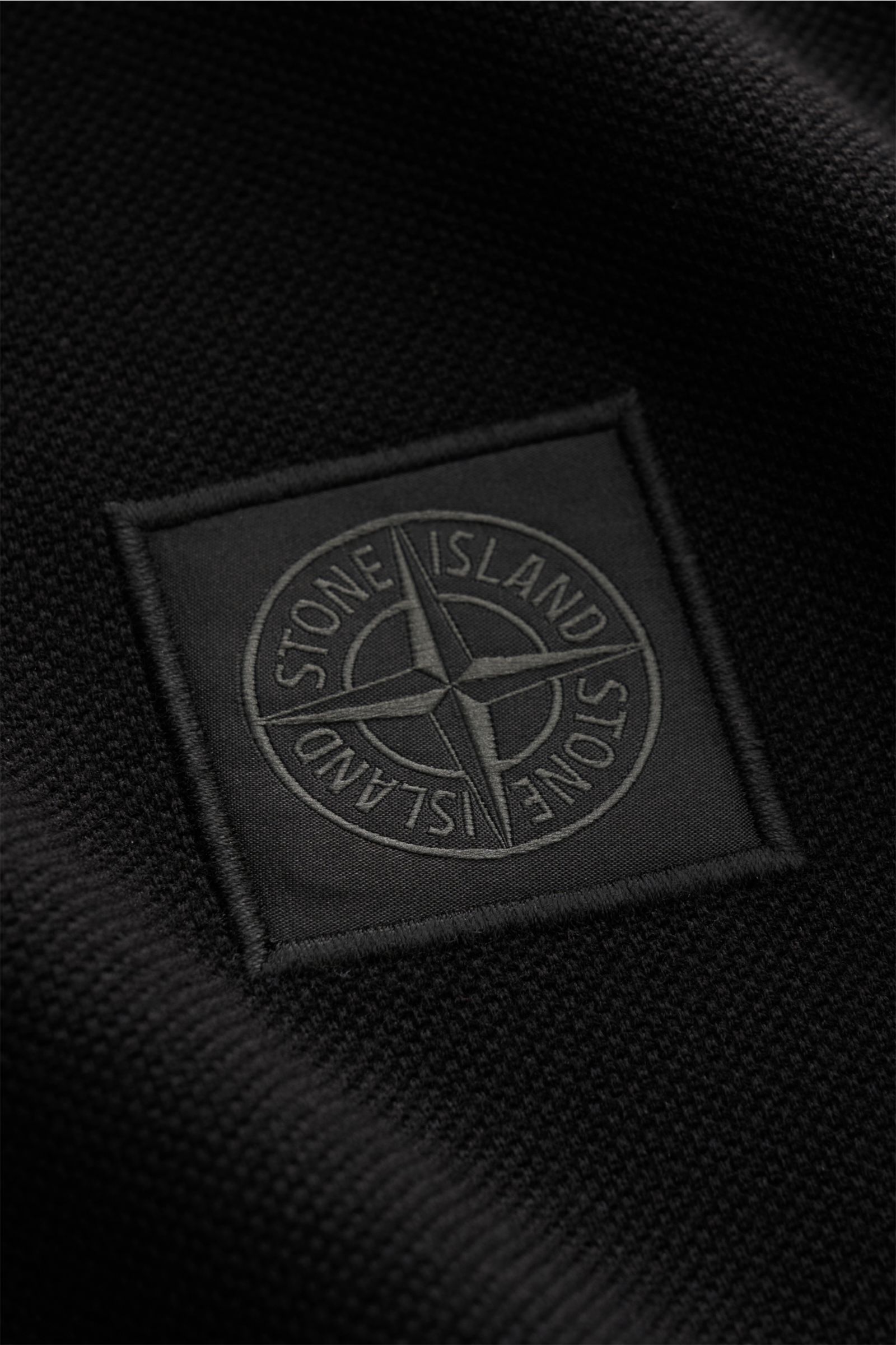 Nahaufnahme des tonalen STONE ISLAND-Weblabels auf dem schwarzen Stone Island Poloshirt 'Ghost Piece' aus Baumwoll-Piqué.