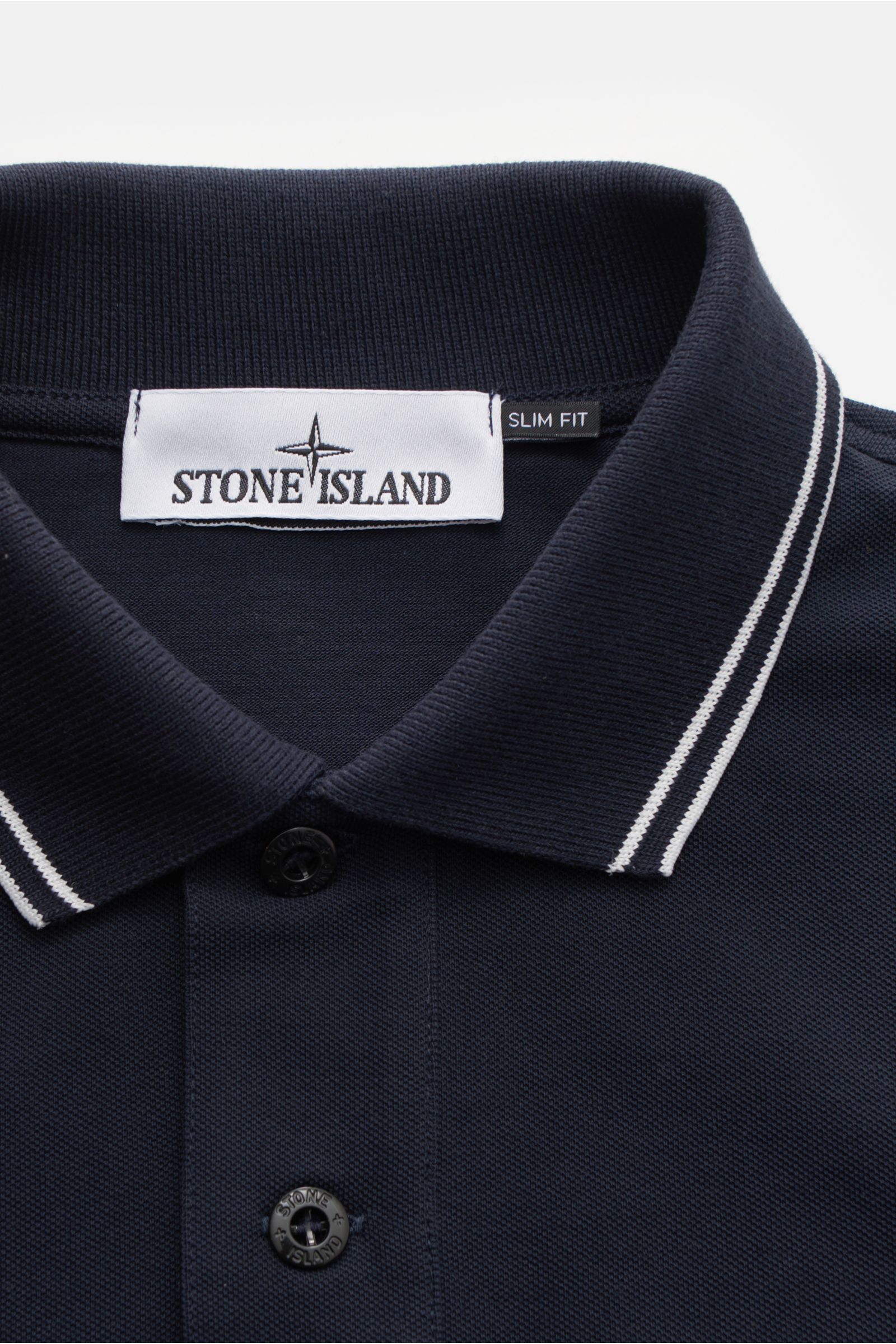 Stone Island Poloshirt navy shown from a close-up top-down perspective, featuring a slim fit piqué cotton fabric with soft texture, narrow white contrast stripes on the collar, typical polo collar, button placket with Stone Island-branded buttons, and ela