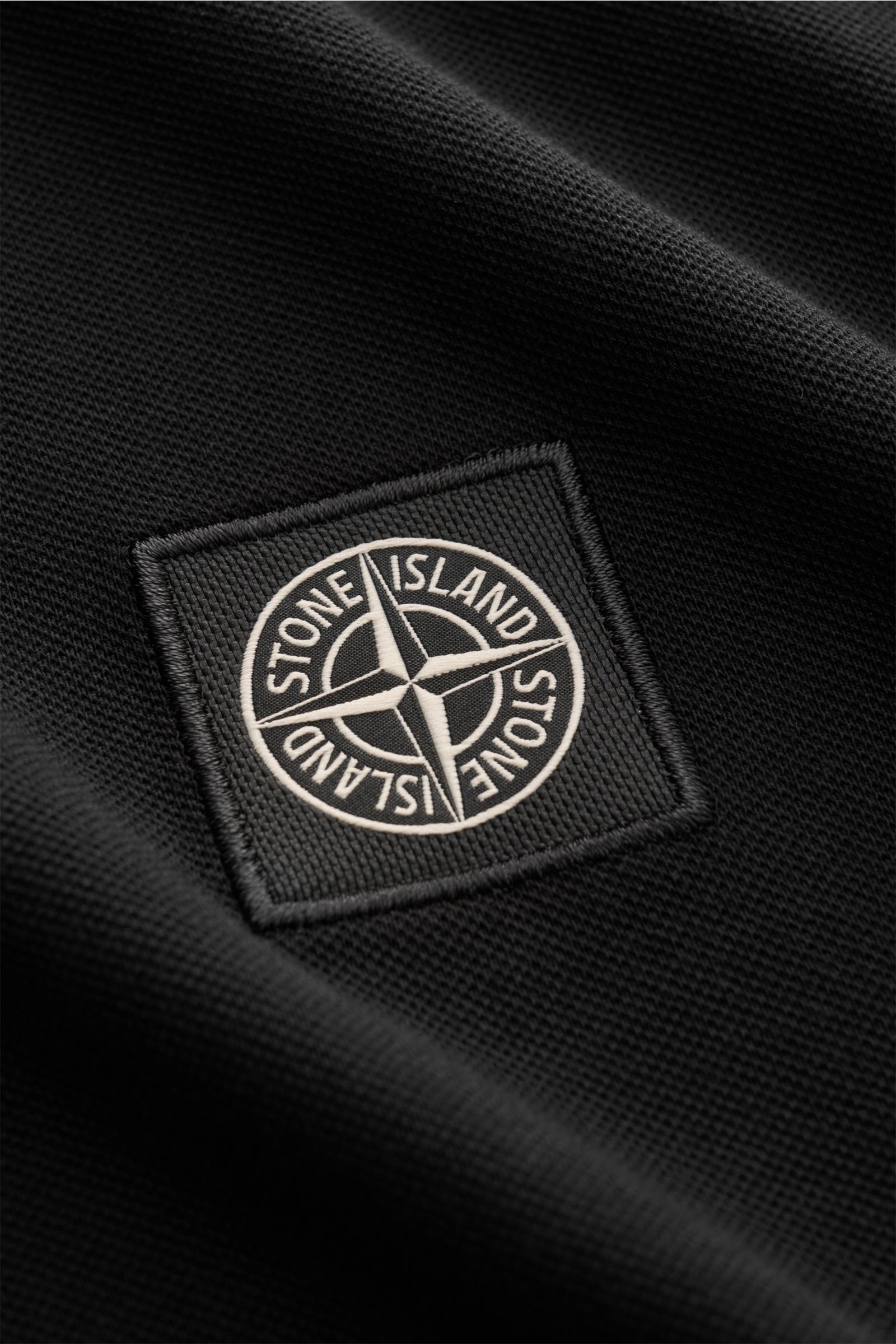 Nahaufnahme des Stone Island Poloshirts schwarz aus elastischem Baumwoll-Piqué mit weichem Griff, sichtbares Weblabel, schmale Kontraststreifen und elastische Ärmelbündchen.