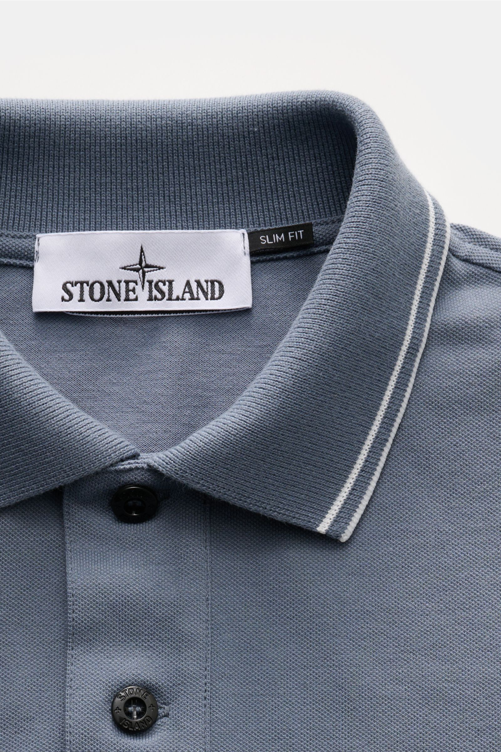 Stone Island Poloshirt graublau close-up from above showing the collar with slim fit label, black buttons, and narrow white contrast stripes on the modern pique cotton fabric. Das moderne Piqué-Poloshirt von STONE ISLAND mit schmalen Kontraststreifen i