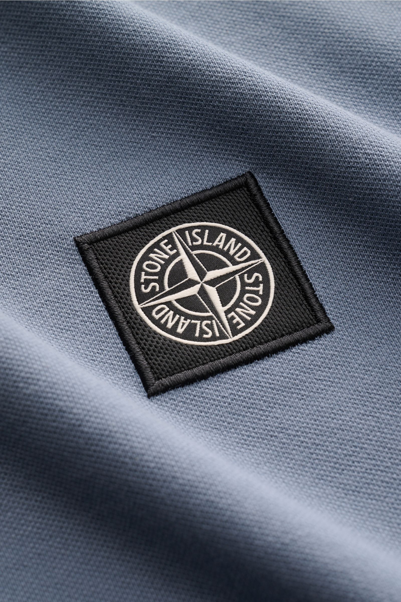 Close-up angled view of the Stone Island Poloshirt graublau showing the black and white Stone Island logo patch on soft, elastic grey-blue piqué cotton fabric.

Description:  
Das moderne Piqué-Poloshirt von STONE ISLAND mit schmalen Kontraststreifen i