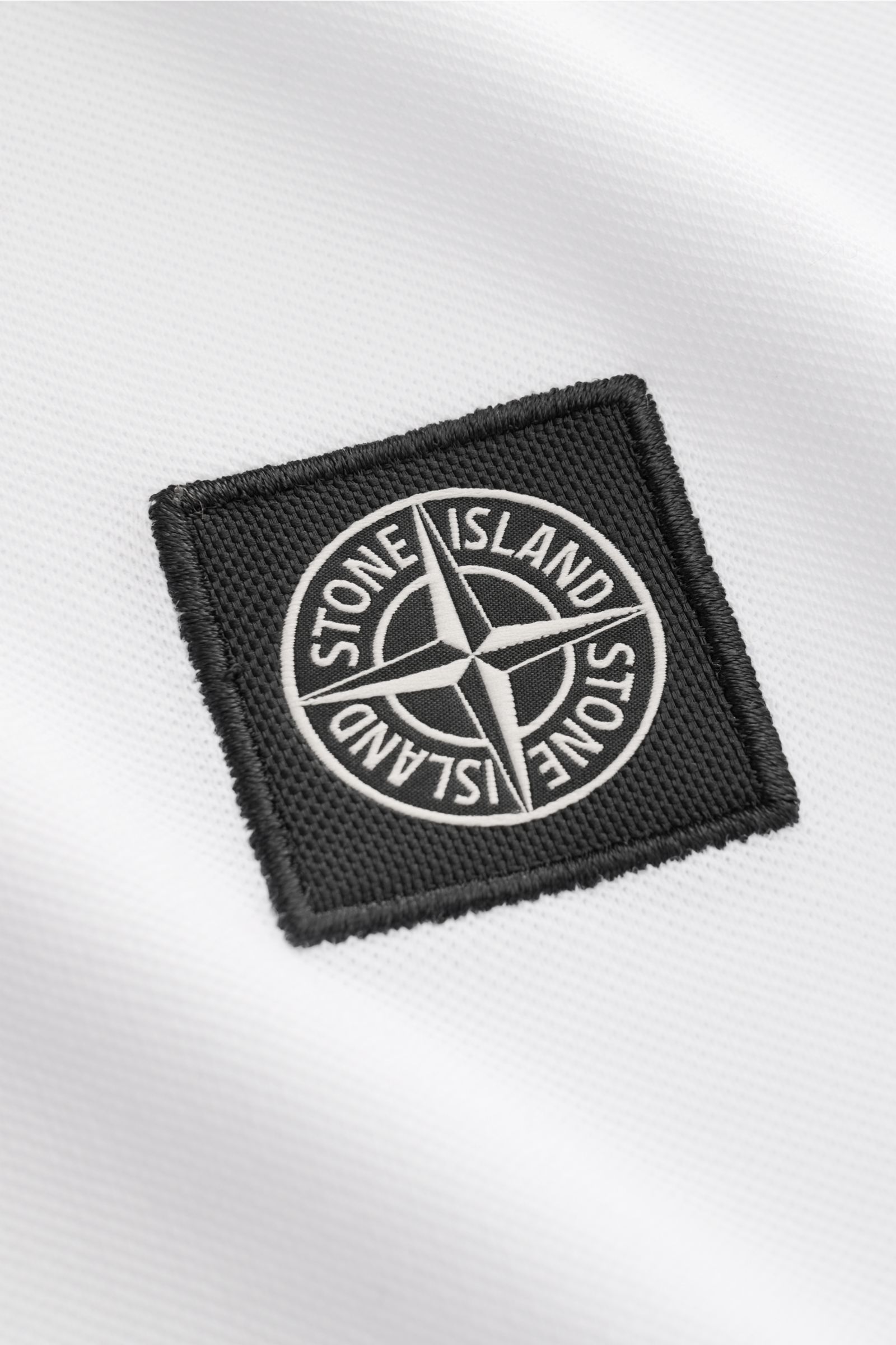 Stone Island Poloshirt weiß aus elastischer Baumwolle in Nahaufnahme, Fokus auf schwarzem Weblabel, Piqué-Struktur sichtbar.