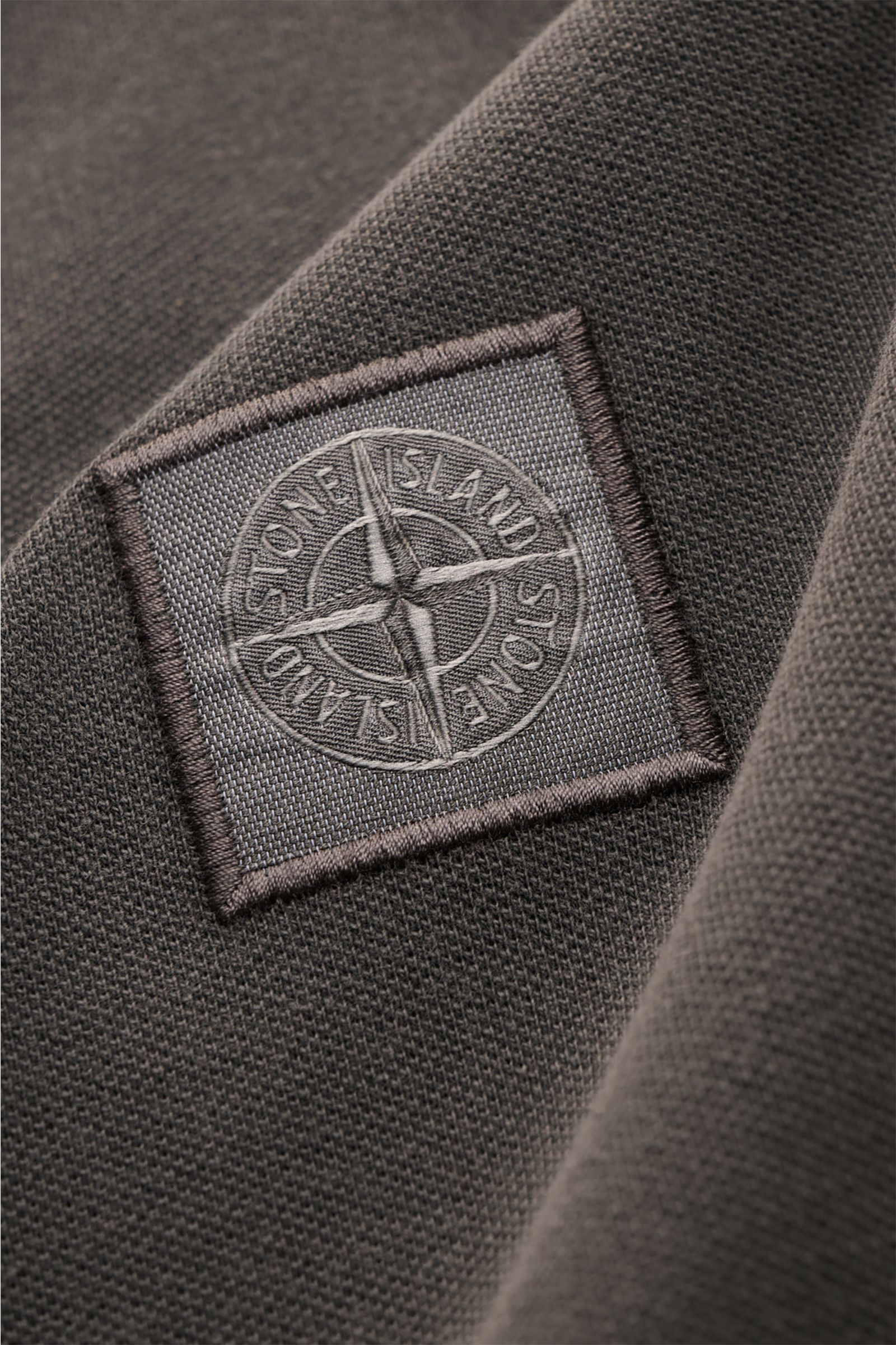 Nahaufnahme des Stone Island Poloshirts dunkelgrau aus elastischer Baumwolle mit STONE ISLAND-Weblabel in seitlicher Perspektive.