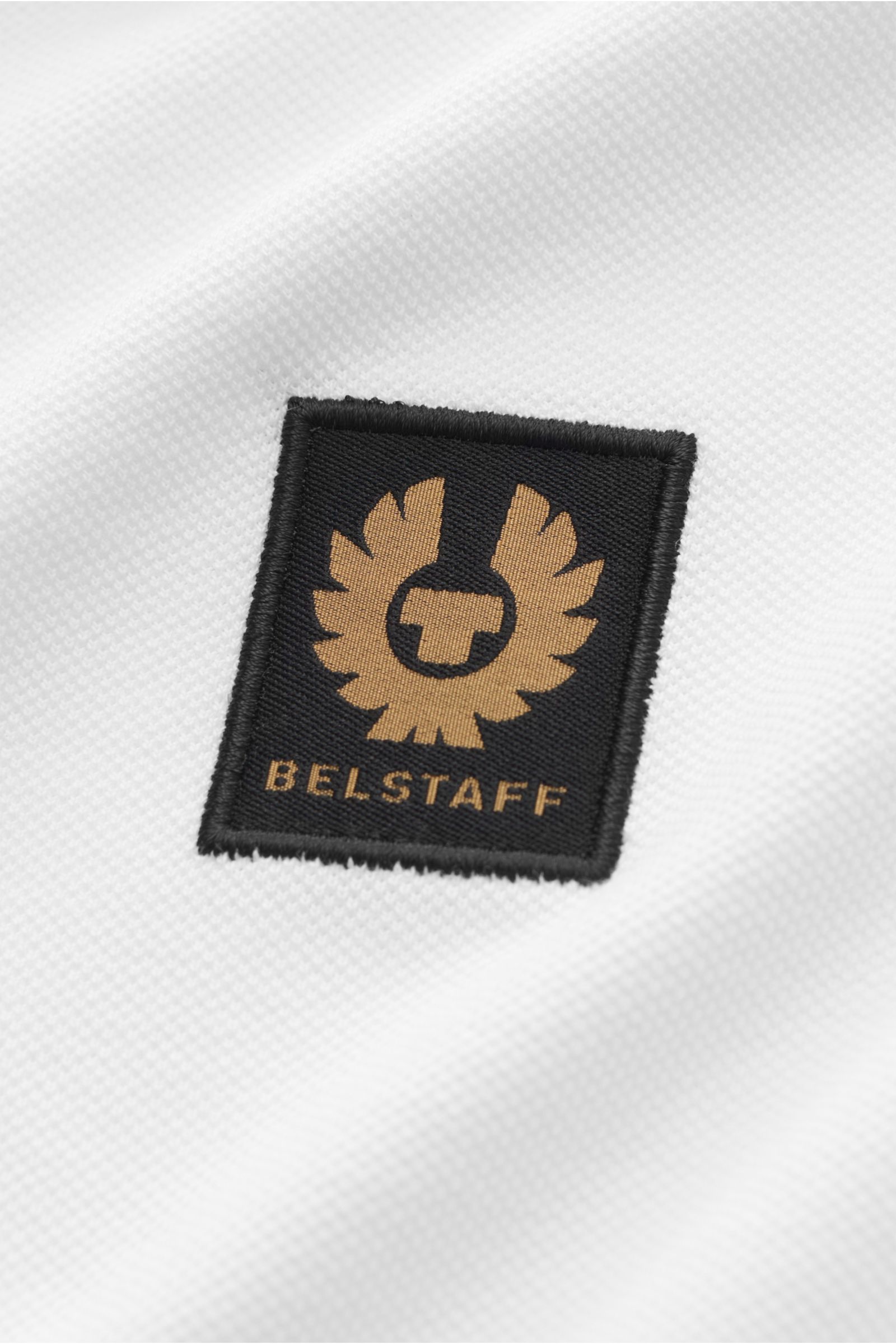 Close-up front view of the Belstaff Poloshirt weiß showing the black and gold BELSTAFF Weblabel on white piqué pure cotton fabric with a slightly textured feel, Slim Fit, two-button placket, classic polo collar, side hem slits, and slightly extended back.