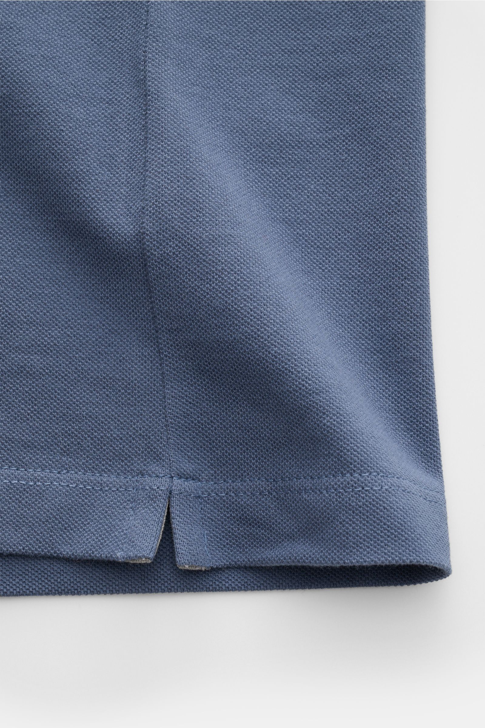 Close-up image from the side showing the hem and side slit of a blue fabric polo shirt, highlighting the piqué texture and stitching details of the Brunello Cucinelli Poloshirt weiß. Cleanes Essential erster Wahl: Liebhaber der Luxusmarke BRUNELLO CUCI