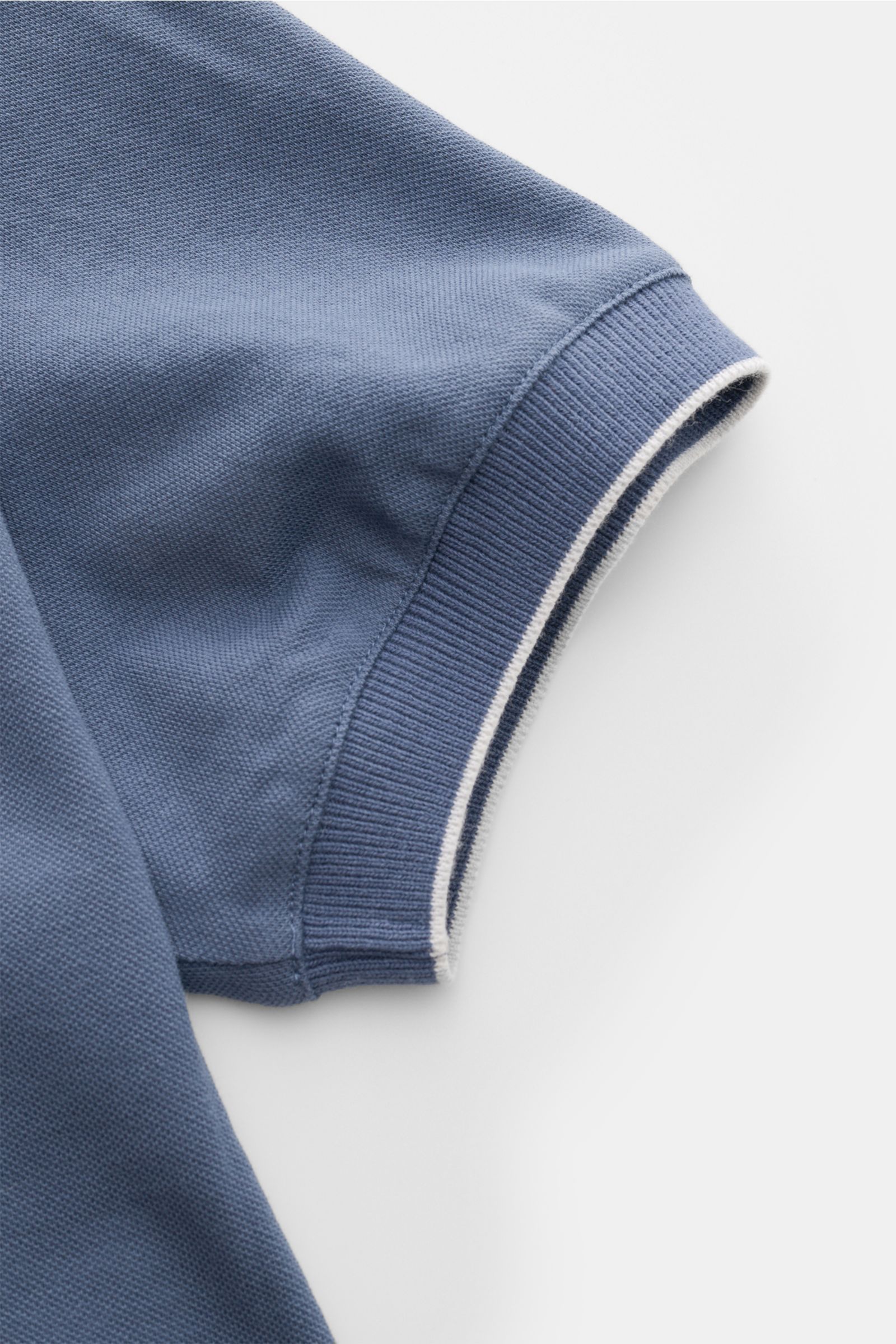 Close-up image of the sleeve of a Brunello Cucinelli Poloshirt weiß in blue, showing the ribbed cuff with a narrow white contrast edge, photographed from above. Cleanes Essential erster Wahl: Liebhaber der Luxusmarke BRUNELLO CUCINELLI sind mit diesem 