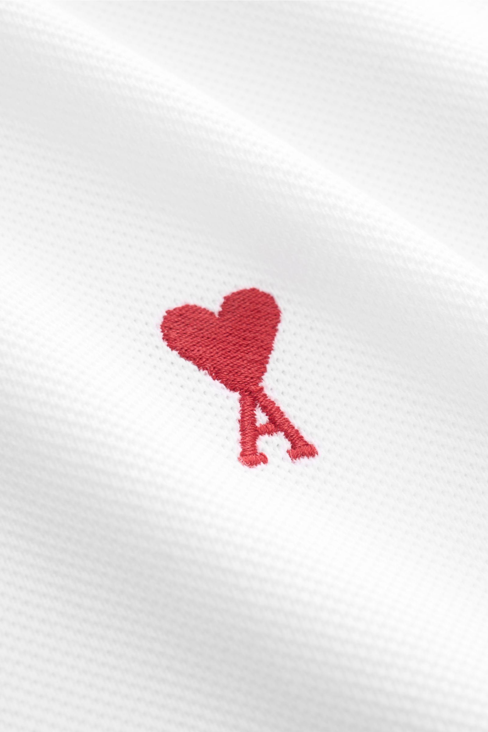 Nahaufnahme des weißen Ami Paris Poloshirts mit rotem „A“-Herz-Stitching aus Piqué-Baumwolle in leichter, griffiger Qualität.