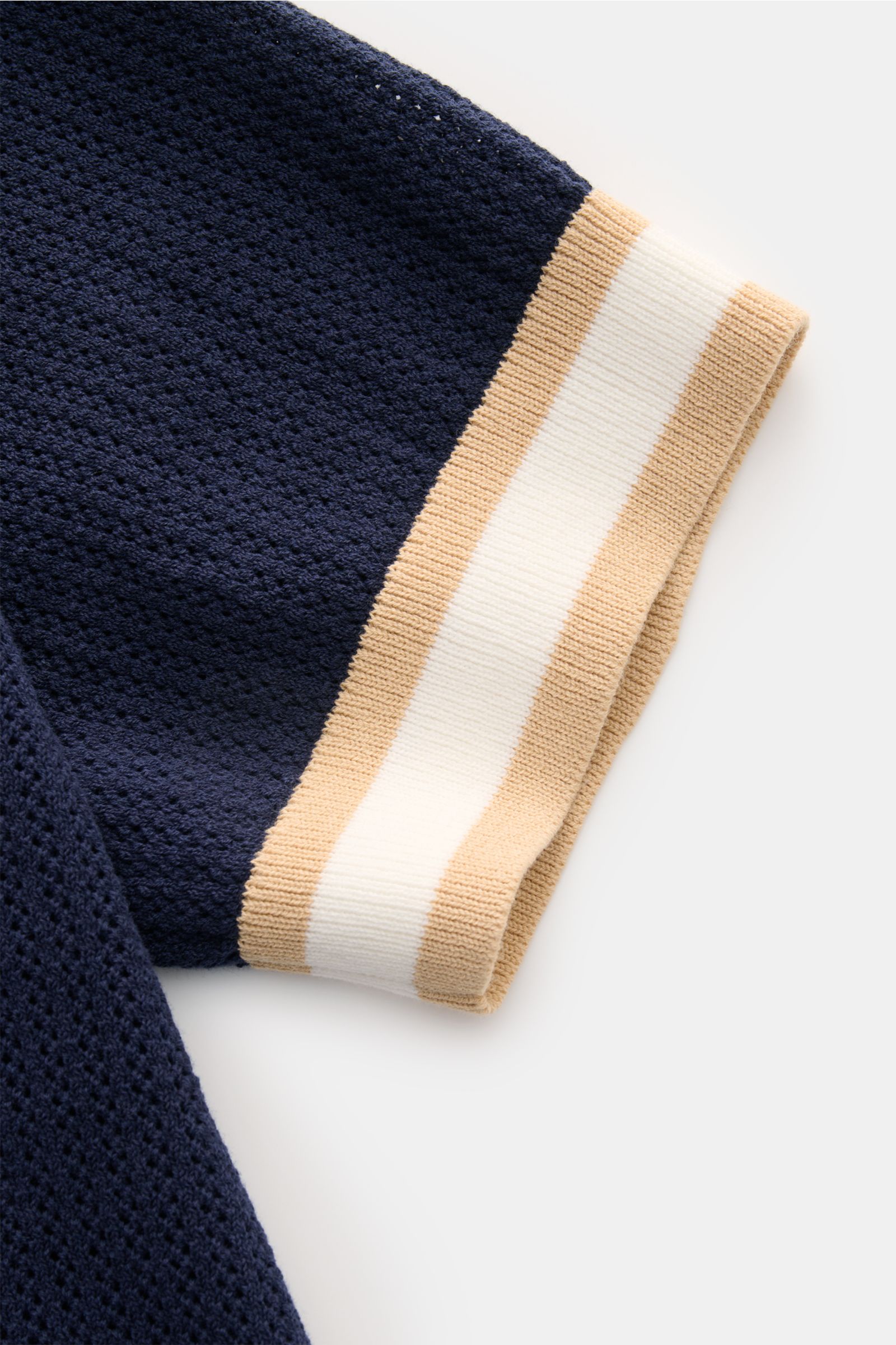 Close-up view of the sleeve of the Ché Kurzarm-Strickpolo 'Quinn' navy/hellbraun, showing navy blue airy knit fabric with beige and white striped ribbed cuff. Genießen Sie die Leichtigkeit des Sommers mit dem luftigen Kurzarm-Strickpolo 'Quinn' von CHÉ. K
