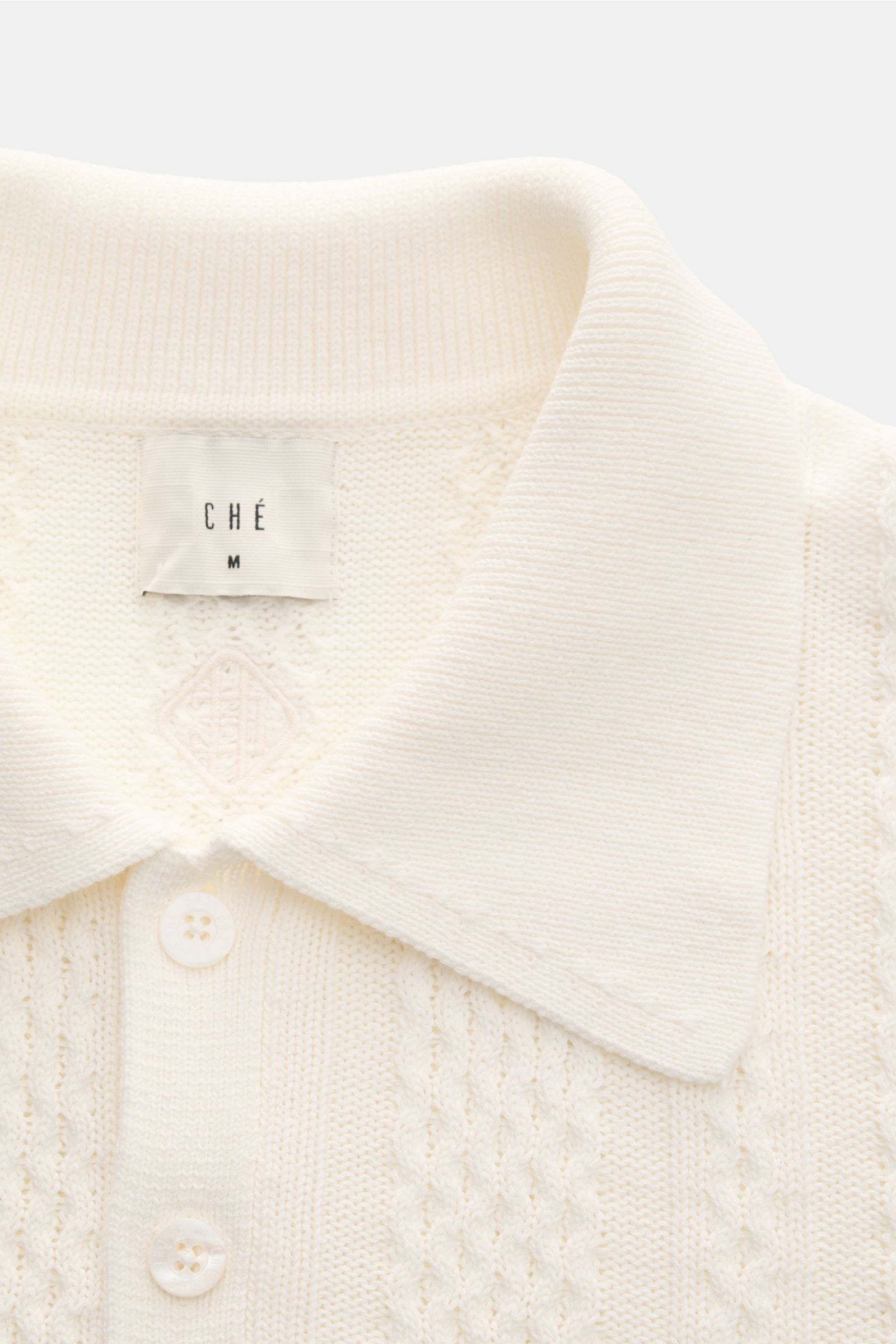 Close-up front view of the Ché Kurzarm-Strickpolo 'Santino' creme, showcasing a heavy quality cream knit polo shirt with an asymmetrical pattern, knitted collar, three-button placket, and tonal CHÉ label. Strickkunst mit Twist: Das 'Santino' Kurzarm-Polos