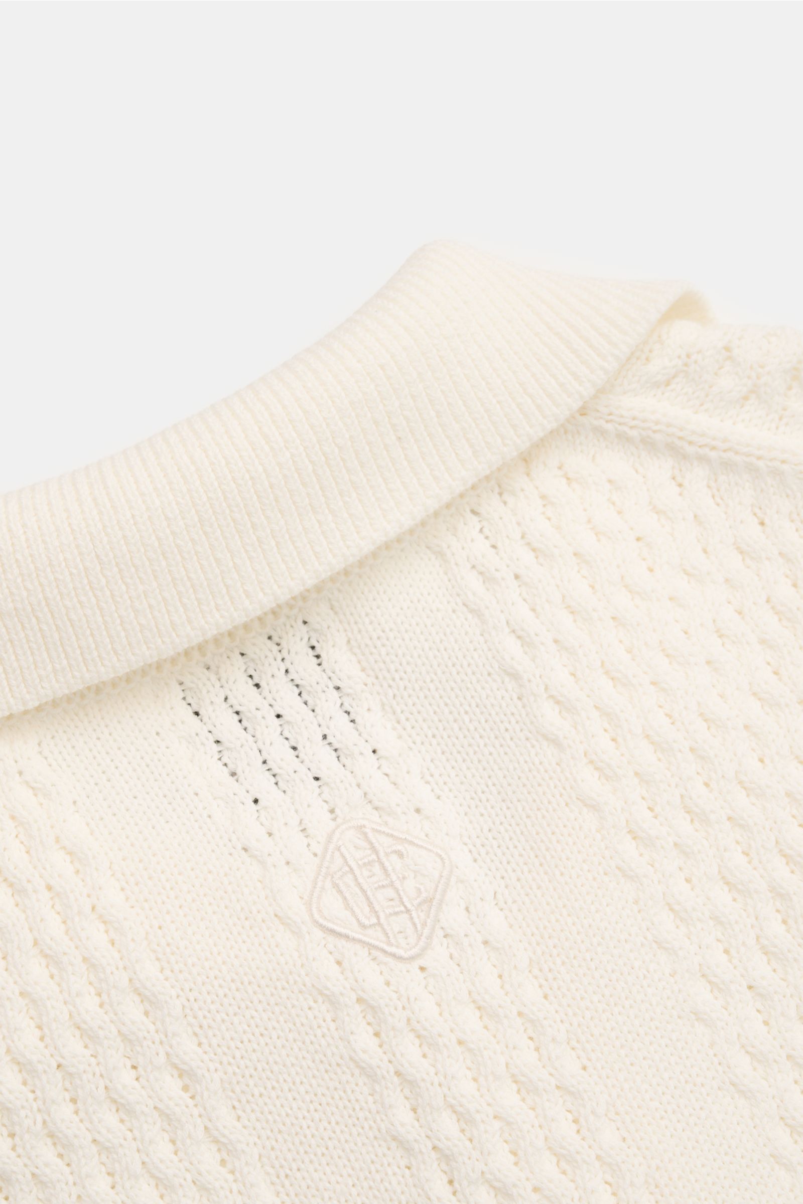 Close-up image of the Ché Kurzarm-Strickpolo 'Santino' creme, showing the knitted polo collar and asymmetrical textured pattern with tonal CHÉ logo from a top-front perspective. Strickkunst mit Twist: Das 'Santino' Kurzarm-Poloshirt von CHÉ überzeugt mit 