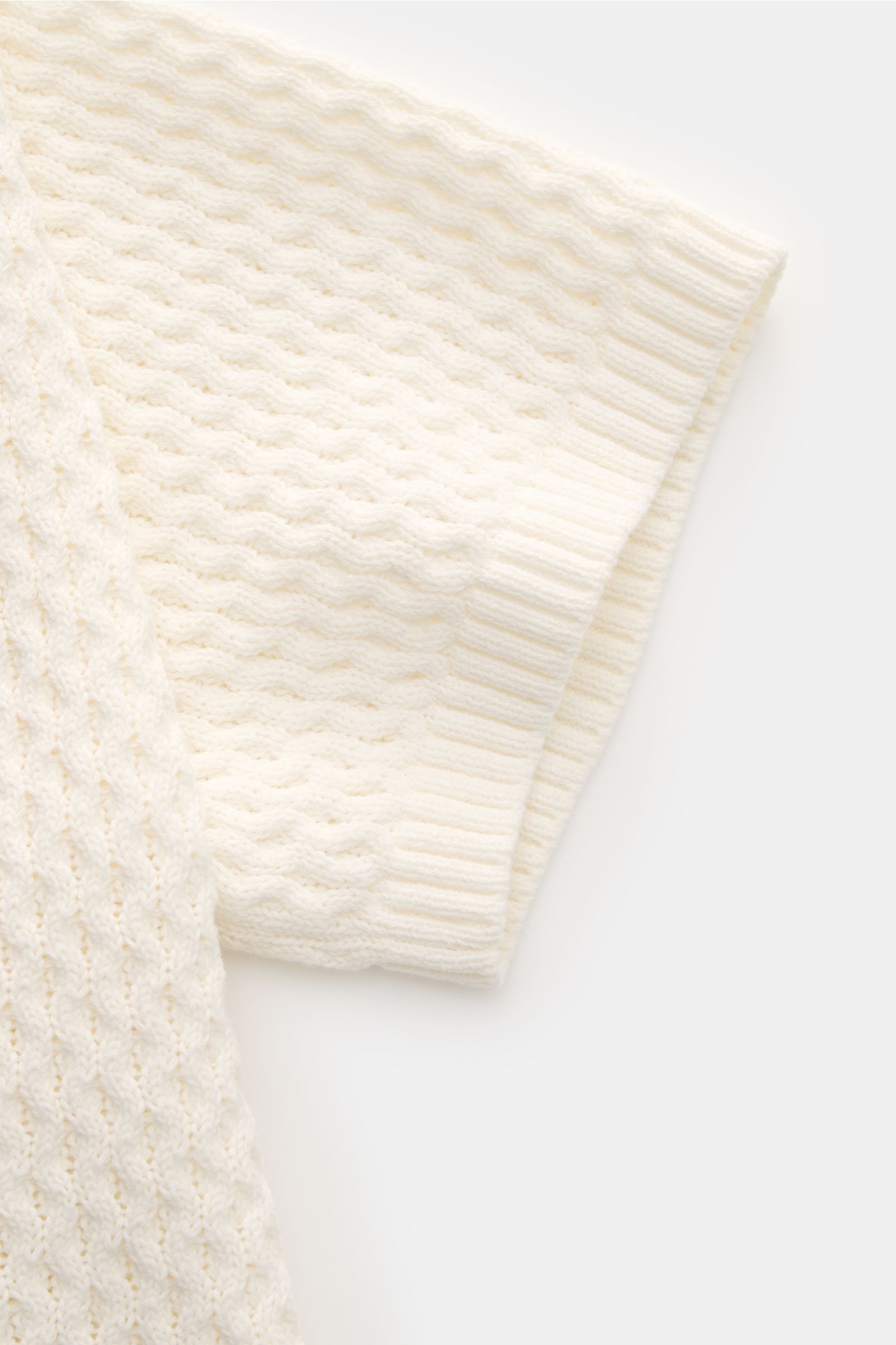 Close-up front view of the sleeve and upper chest area of the Ché Kurzarm-Strickpolo 'Santino' creme, showcasing its coarse asymmetric knit pattern and ribbed cuffs. Strickkunst mit Twist: Das 'Santino' Kurzarm-Poloshirt von CHÉ überzeugt mit einem groben