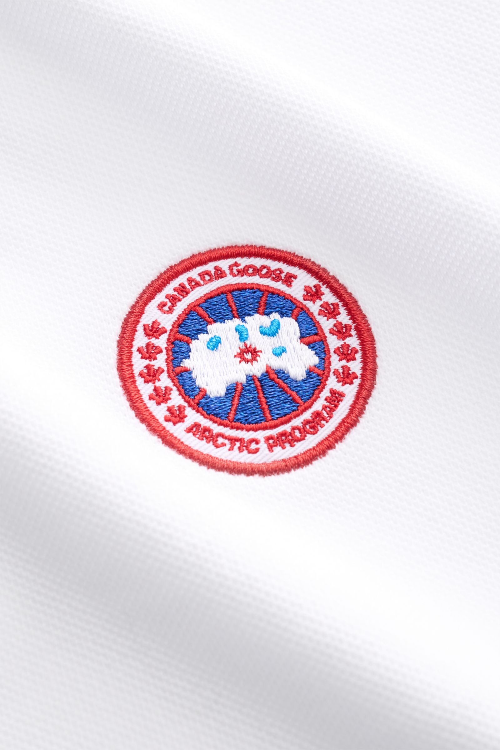 Canada Goose Poloshirt 'Beckley' weiß shown in close-up from a front angle, highlighting the embroidered CANADA GOOSE patch on white cotton piqué fabric. Das klassische Poloshirt 'Beckley' ist ein Must-have in der sportiven Herrengarderobe: Aus Baumwol