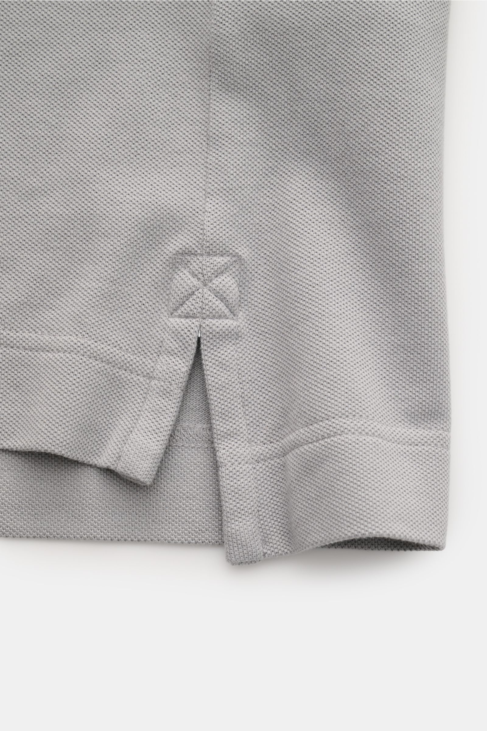 Close-up image of the side hem of the Canada Goose Poloshirt 'Beckley' grau, showing the textured cotton piqué fabric, side slit with reinforced stitching, and flexible cuff detail. The product is photographed from a side perspective. Das klassische Po