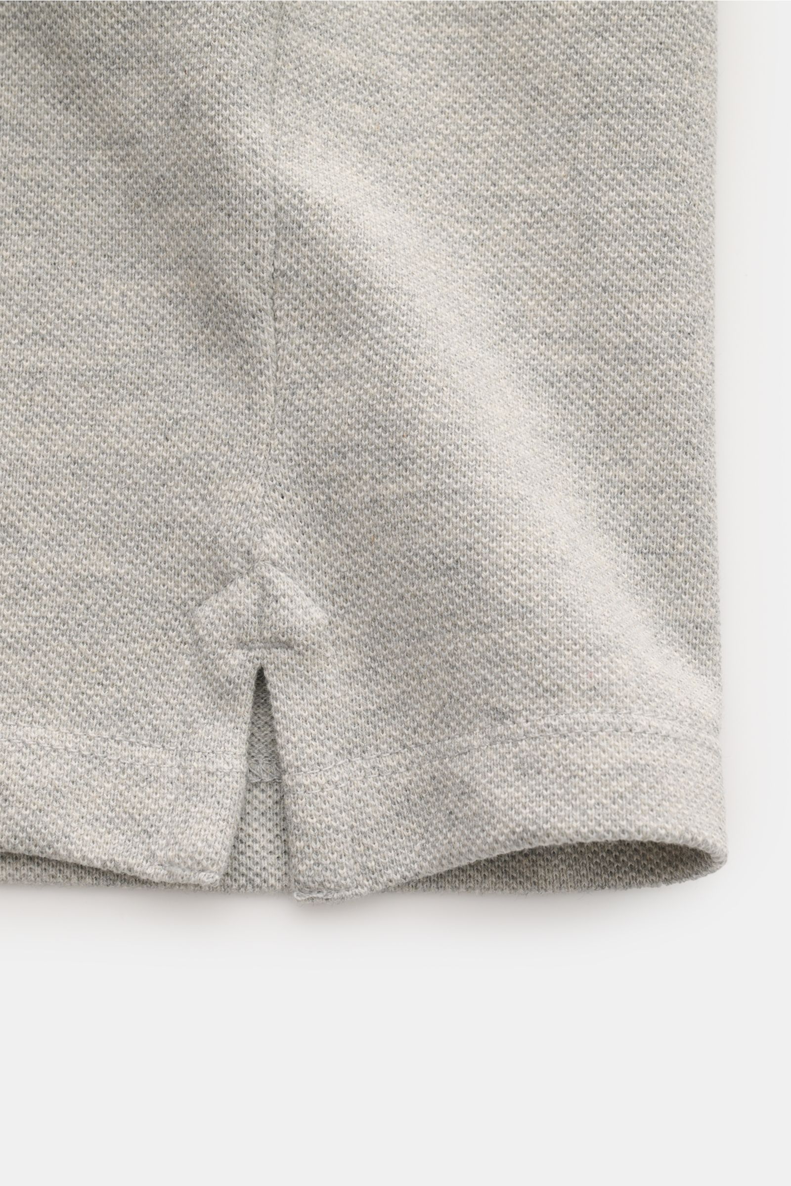 Close-up of the hem detail of the Ami Paris Poloshirt grau meliert, showing the fine piqué structure, organic cotton jersey, elastic sleeve cuff, short side slit, and subtle 'A' logo stitching, photographed from a top-front perspective. 

Ein schöner K