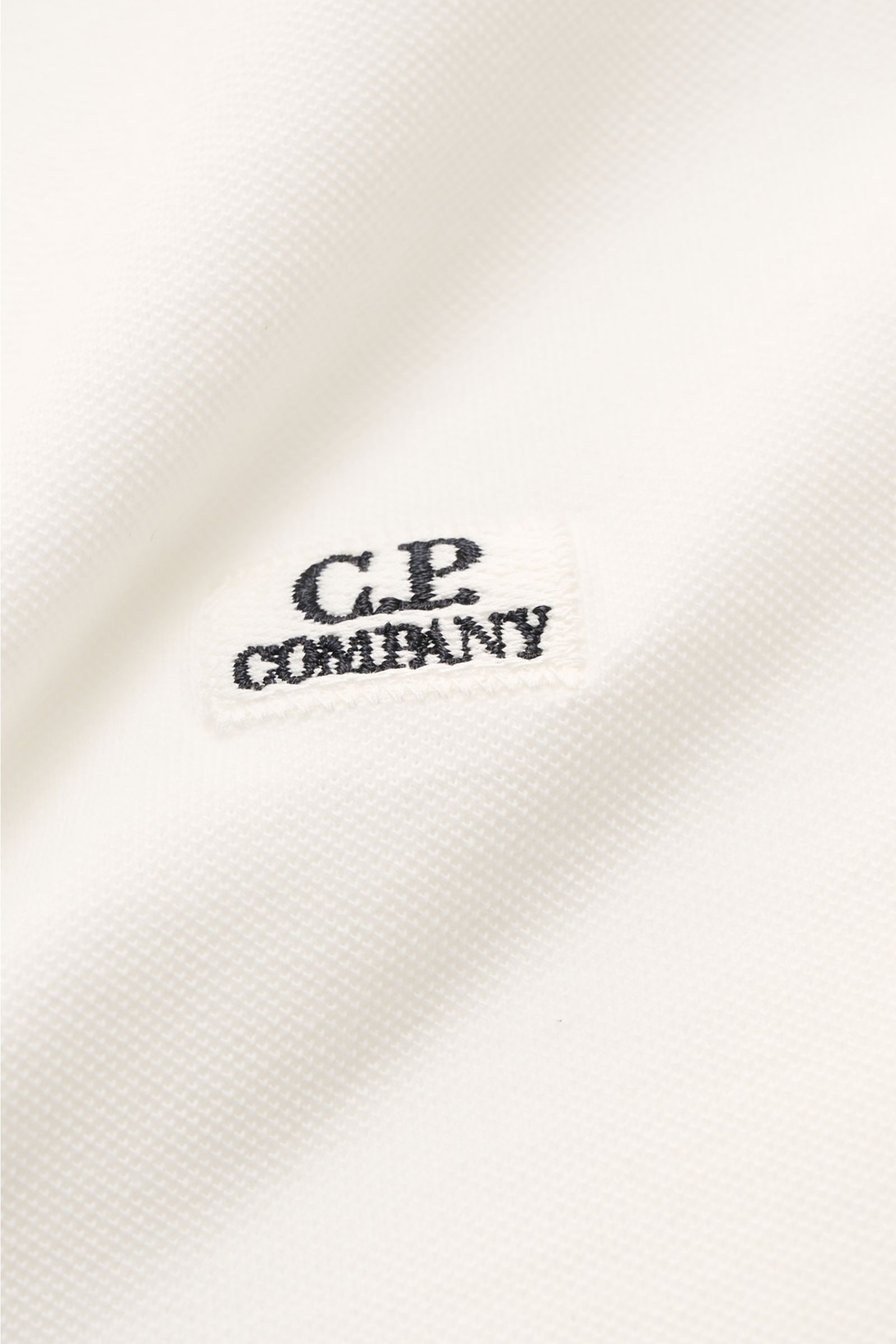 Nahaufnahme des C.P. Company Poloshirts weiß aus Baumwoll-Piqué mit feiner Struktur, kleinem Weblabel und pigmentgefärbter Oberfläche.
