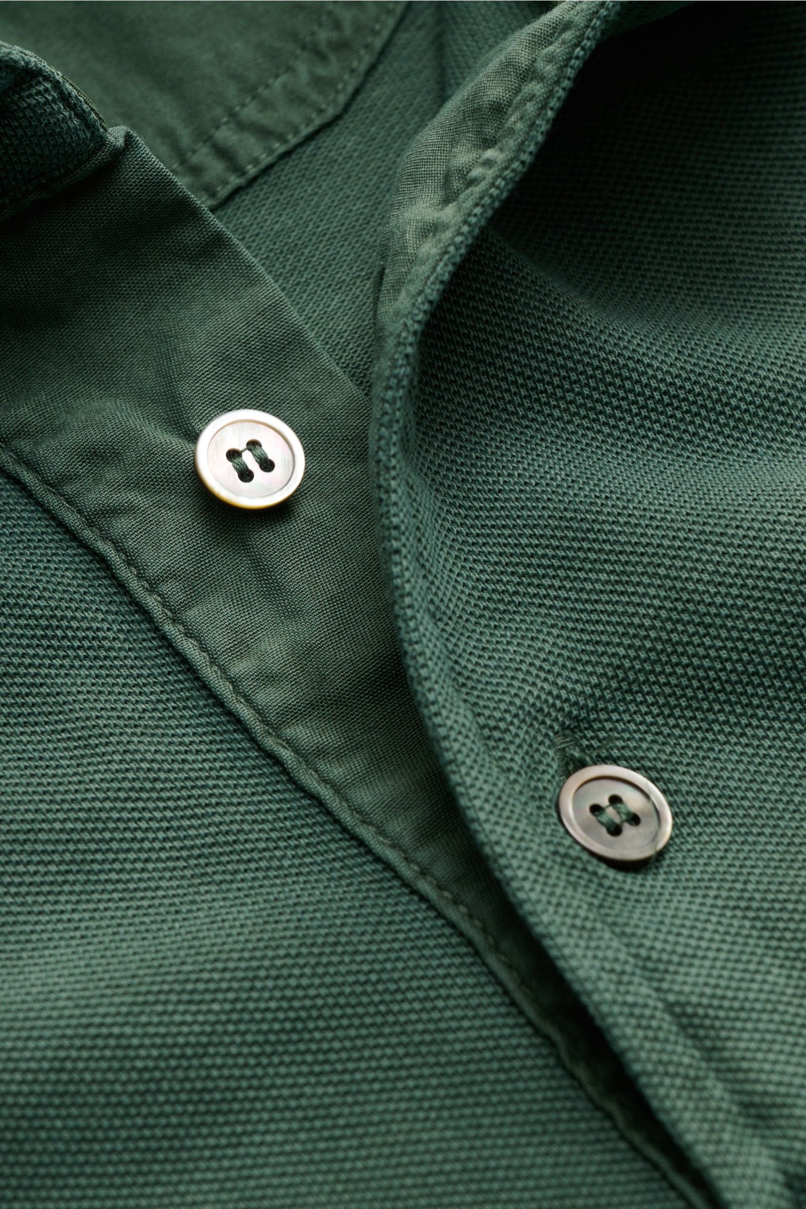 Nahaufnahme des Della Ciana Poloshirts dunkelgrün aus stückgefärbter Baumwolle, feine Piqué-Struktur, Knopfleiste, Perspektive von oben.