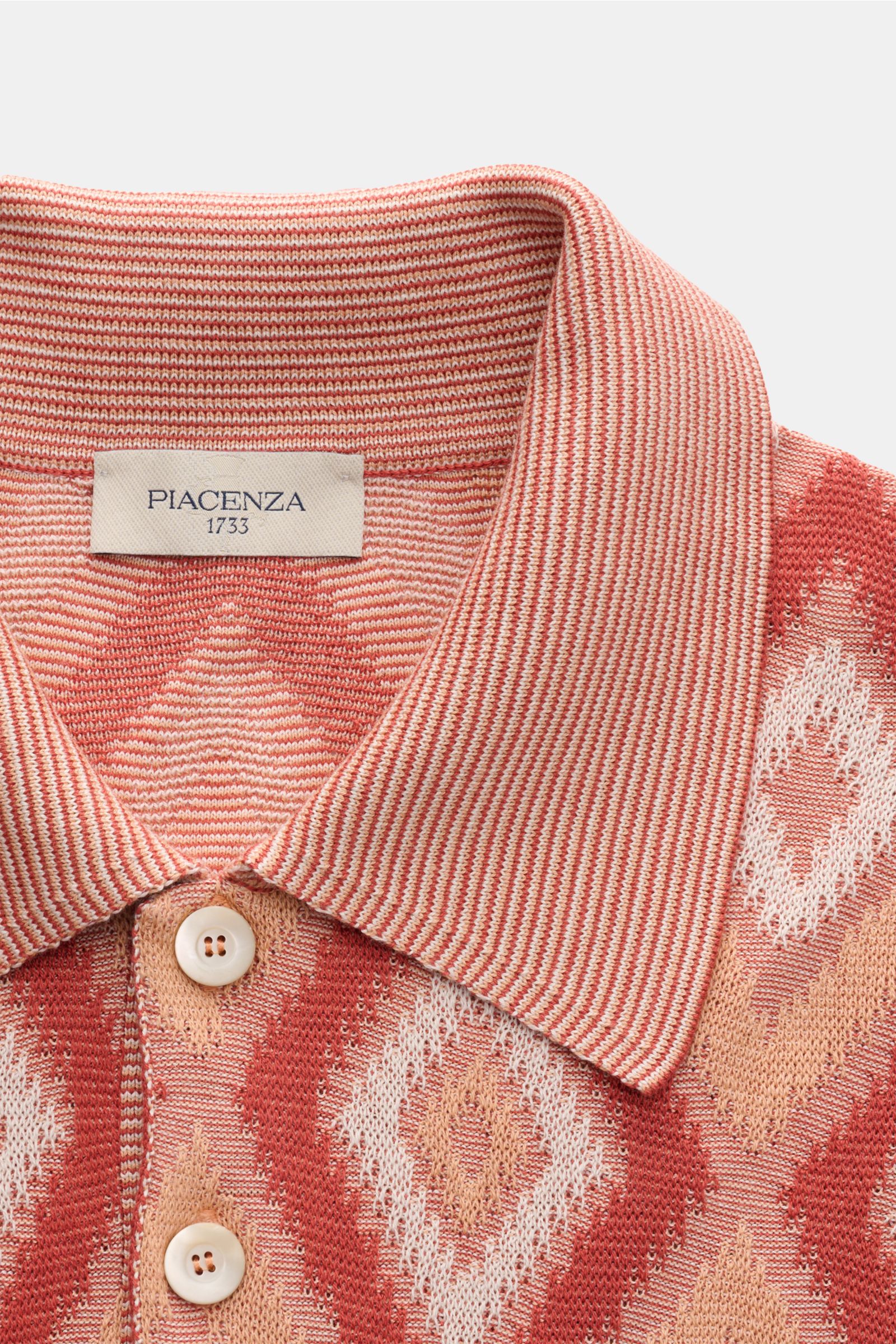 Close-up, top-down view of the Piacenza 1733 Kurzarm-Strickpolo rot/apricot/offwhite gemustert, showing its knitted polo collar, three-button placket, and airy diamond pattern in red, apricot, and offwhite. Ein Hauch von Luxus: Dieses Kurzarm-Strickpol