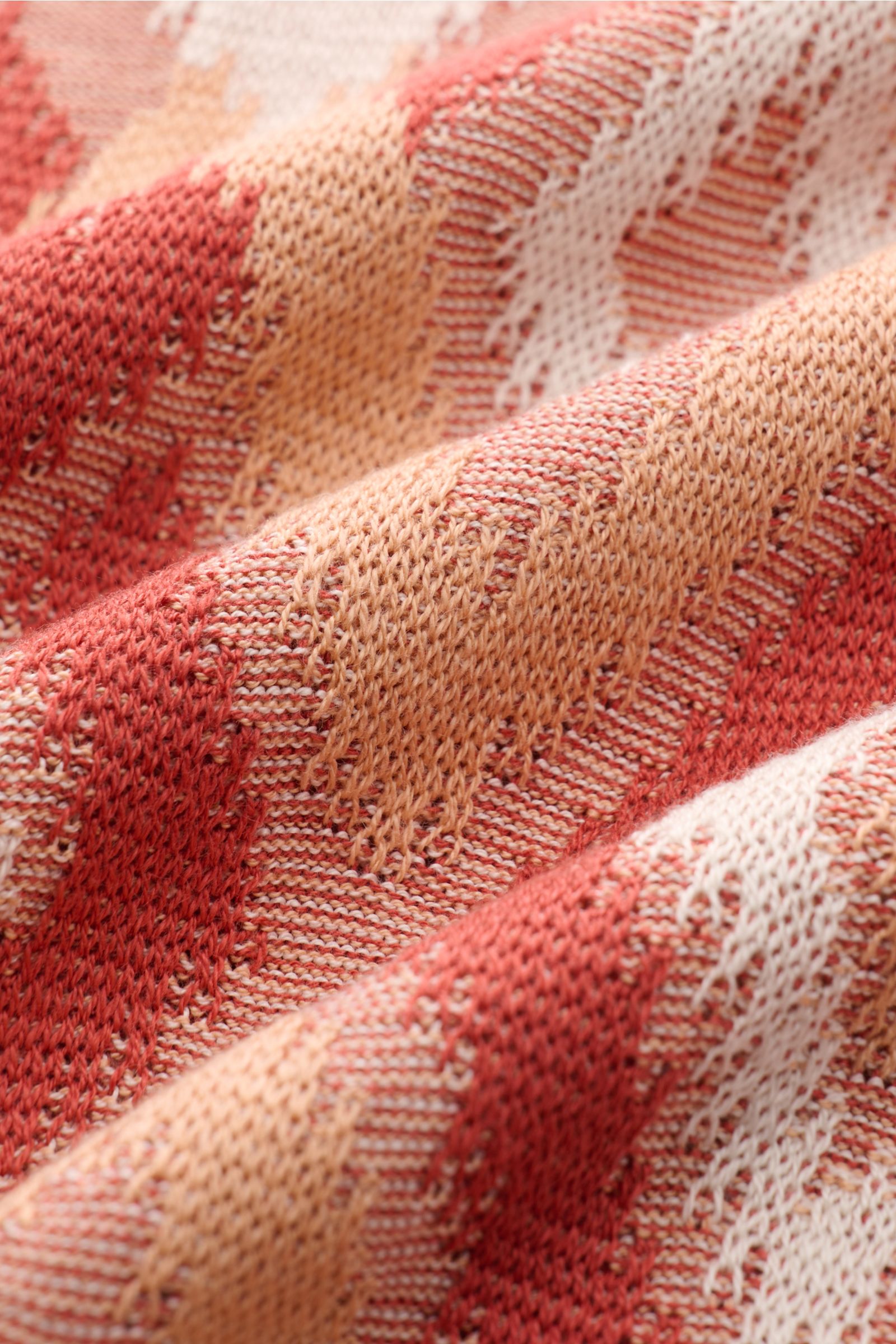 Close-up photo from a side angle of the Piacenza 1733 Kurzarm-Strickpolo rot/apricot/offwhite gemustert, showcasing its lightweight knit fabric with a breathable pattern and contrasting diamond design. Ein Hauch von Luxus: Dieses Kurzarm-Strickpolo von PI