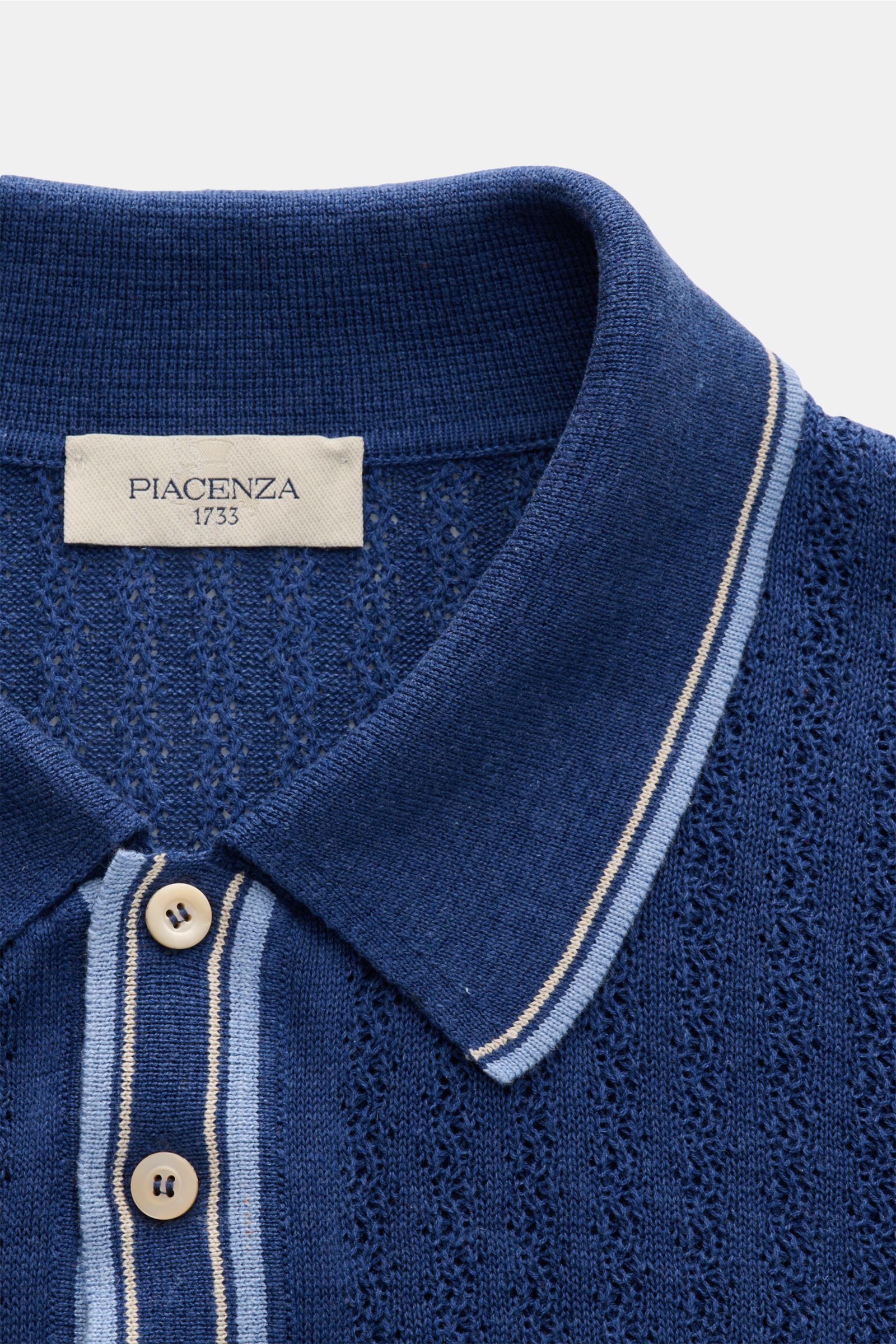 Close-up, top-front view of the Piacenza 1733 Kurzarm-Strickpolo dunkelblau showing its dark blue silk-linen blend, soft texture, striped structured knit, classic polo collar, and button placket.

Eyecatcher in der Sommersaison: Das Kurzarm-Strickpolo 