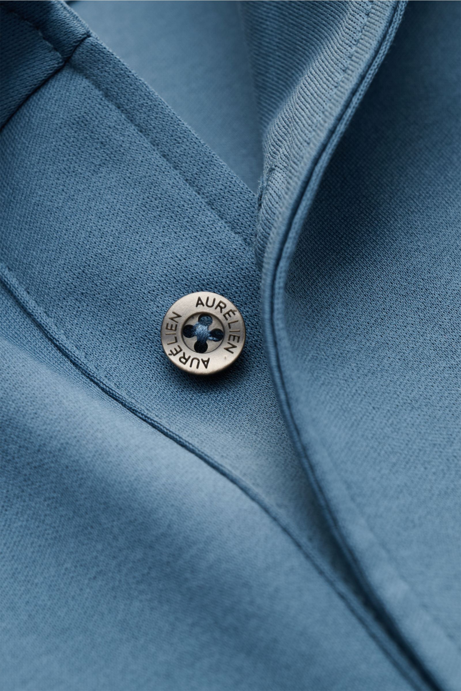 Close-up, top-down view of the Aurélien Jersey-Poloshirt petrol showing a metal button with 'AURÉLIEN' engraved, made of pure cotton with a smooth, cool touch. Vielseitiges Basic für den Frühling und Sommer: Das Jersey-Poloshirt von AURÉLIEN verbindet lan