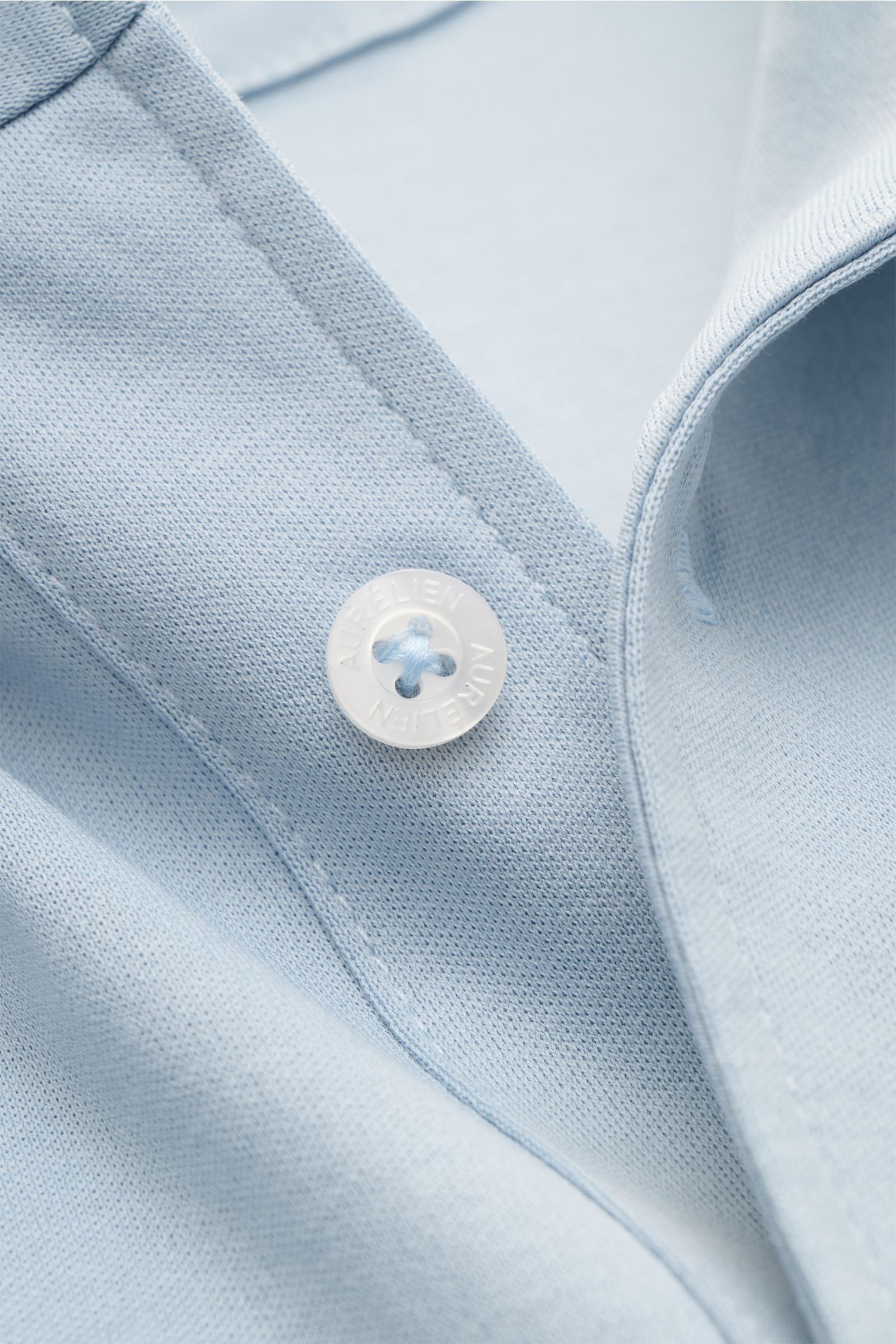 Close-up front view of the Aurélien Jersey-Poloshirt hellblau showing the light blue fine cotton fabric, a white button with the Aurélien logo, and detailed stitching. Vielseitiges Basic für den Frühling und Sommer: Das Jersey-Poloshirt von AURÉLIEN verbi