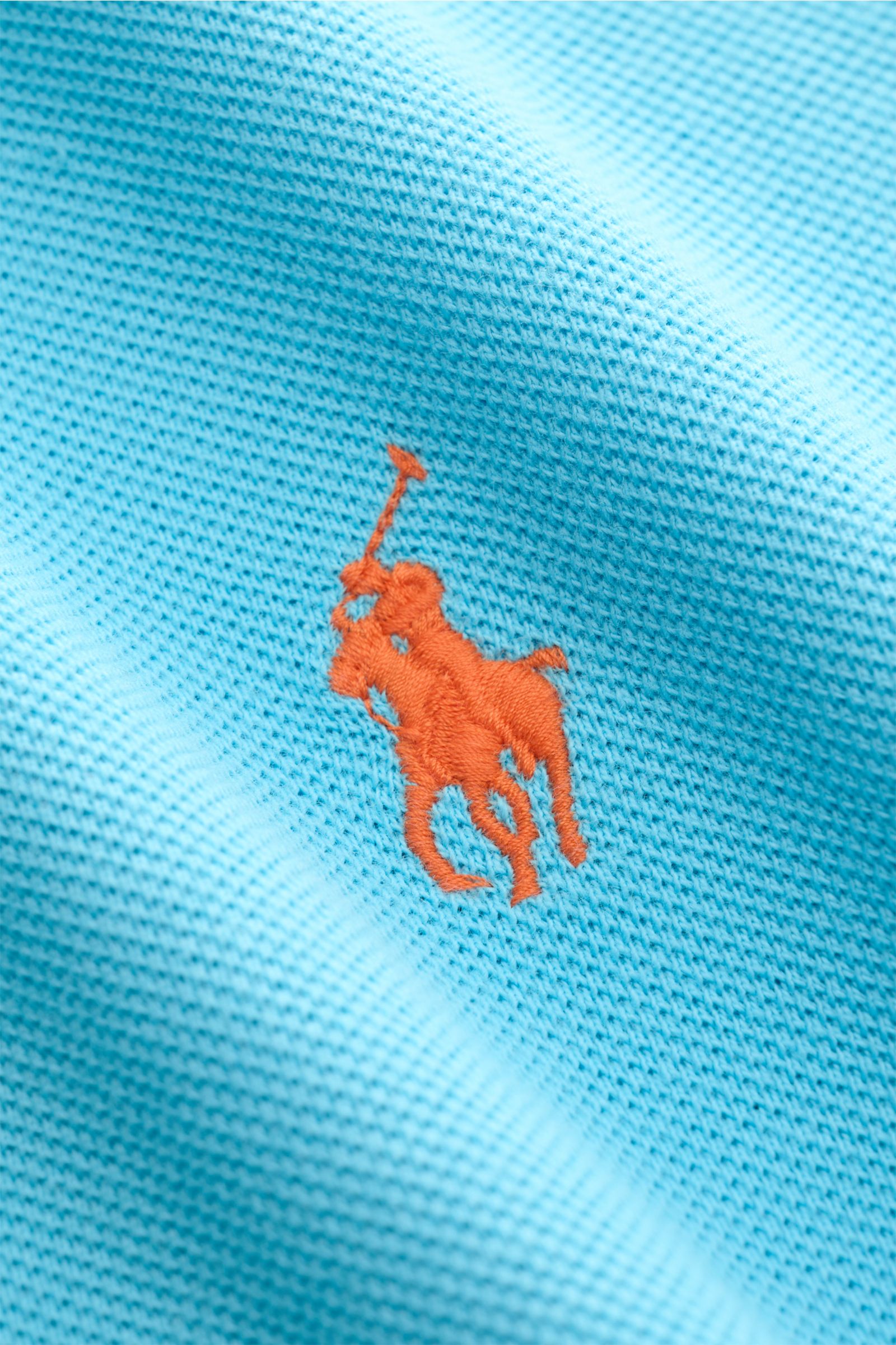 Nahaufnahme aus schräger Perspektive auf das orangefarbene Poloreiter-Stitching des Polo Ralph Lauren Poloshirts azurblau aus reiner Baumwolle mit feiner Piqué-Struktur.