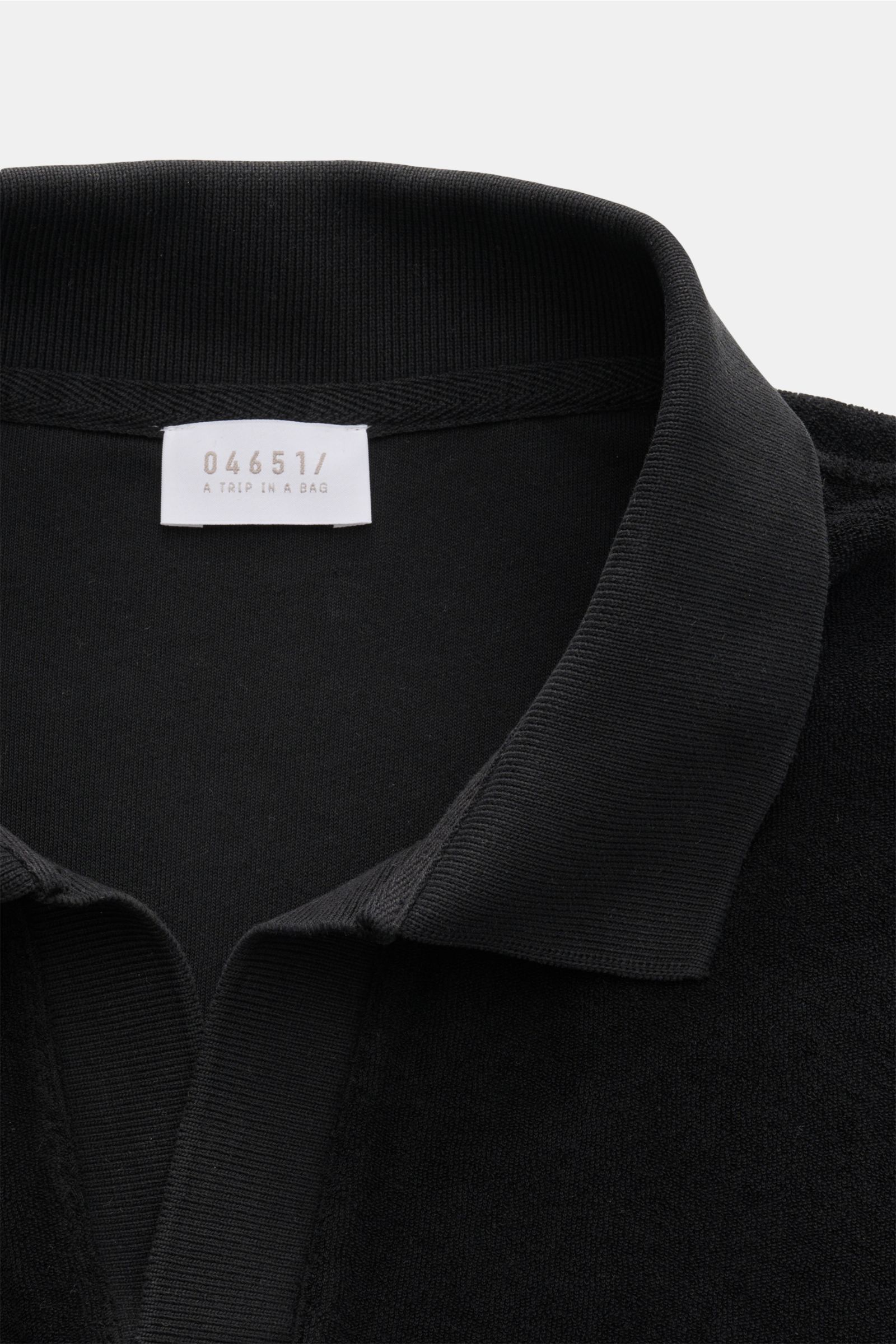 Schwarz fotografierte Nahaufnahme aus schräger Frontperspektive des 04651/ A trip in a bag Frottee-Poloshirt 'Terry Johnny' mit Polokragen, offener Kragenleiste, weichem Baumwoll-Frottee und kleinem Weblabel in der Seitennaht.