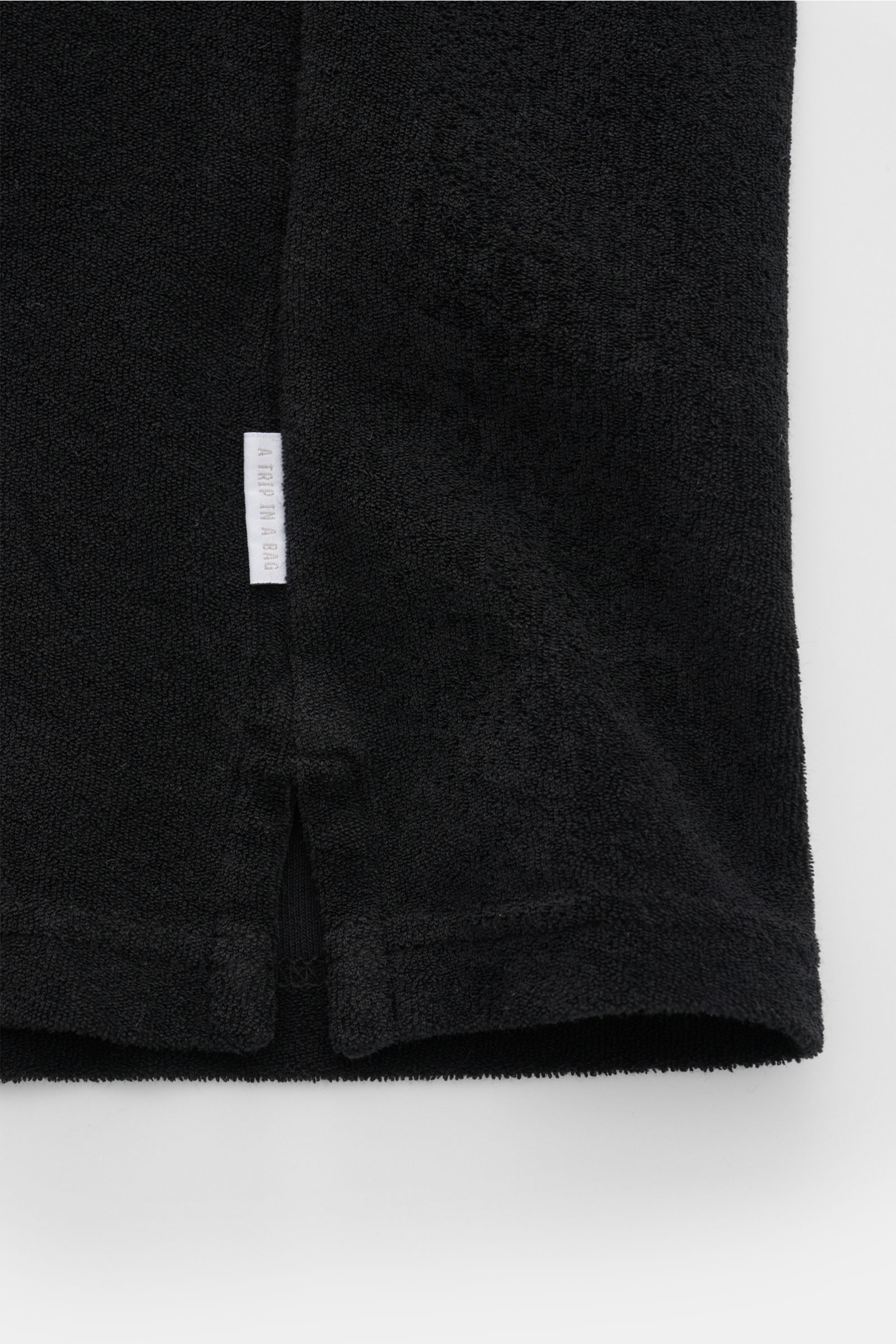 Schwarzes 04651/ A trip in a bag Frottee-Poloshirt 'Terry Johnny' in Regular Fit, Detailansicht Saum mit kurzem Schlitz und kleinem Weblabel.