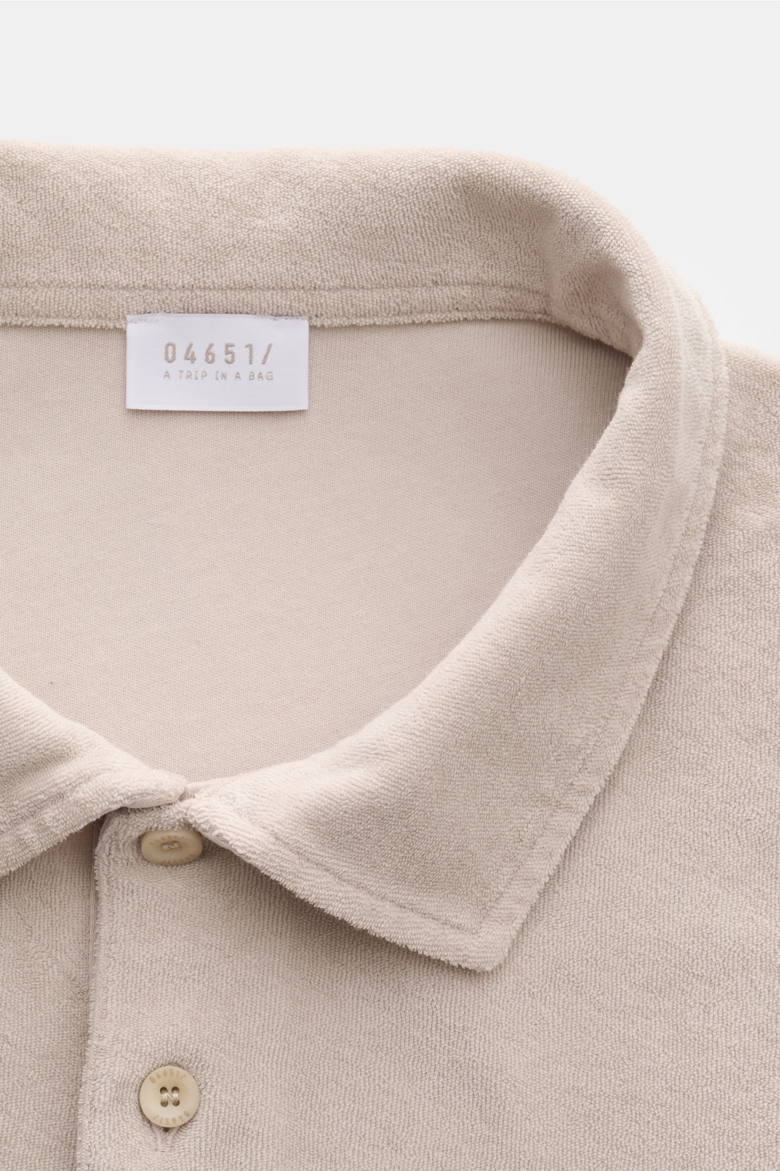 Nahaufnahme des beigen 04651/ A trip in a bag Frottee-Poloshirts 'Terry Polo' aus reiner Baumwolle mit Polokragen, Knopfleiste, weichem Griff und kleinem Weblabel.