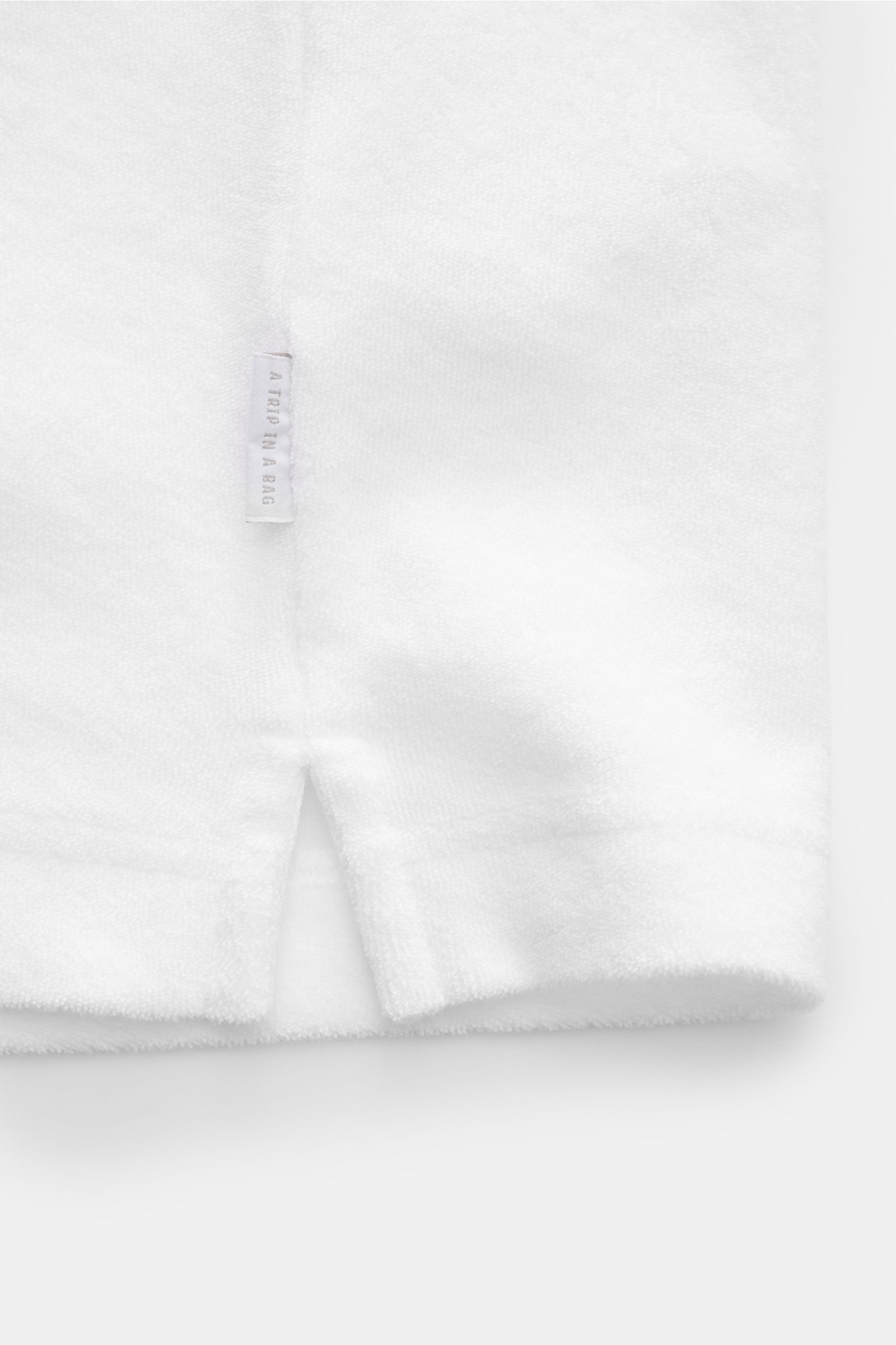 Weiße Detailaufnahme von 04651/ A trip in a bag Frottee-Poloshirt 'Terry Polo' mit kurzem Saumschlitz, kleinem Weblabel, weichem Baumwollstoff.