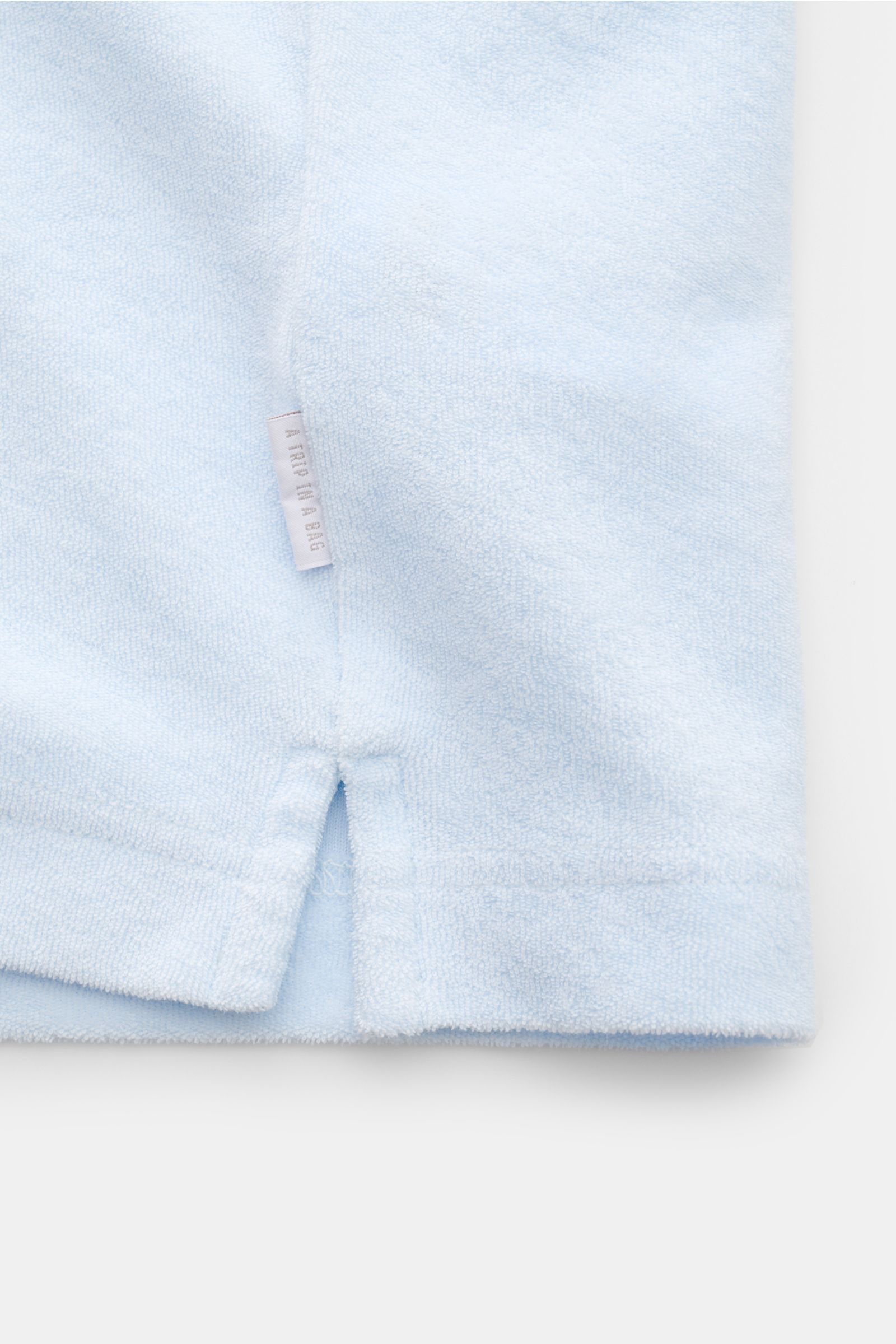 Hellblauer Detailblick auf den unteren Saum des 04651/ A trip in a bag Frottee-Poloshirts 'Terry Polo' mit kurzem Saumschlitz, Weblabel und weichem Baumwollfrottee.