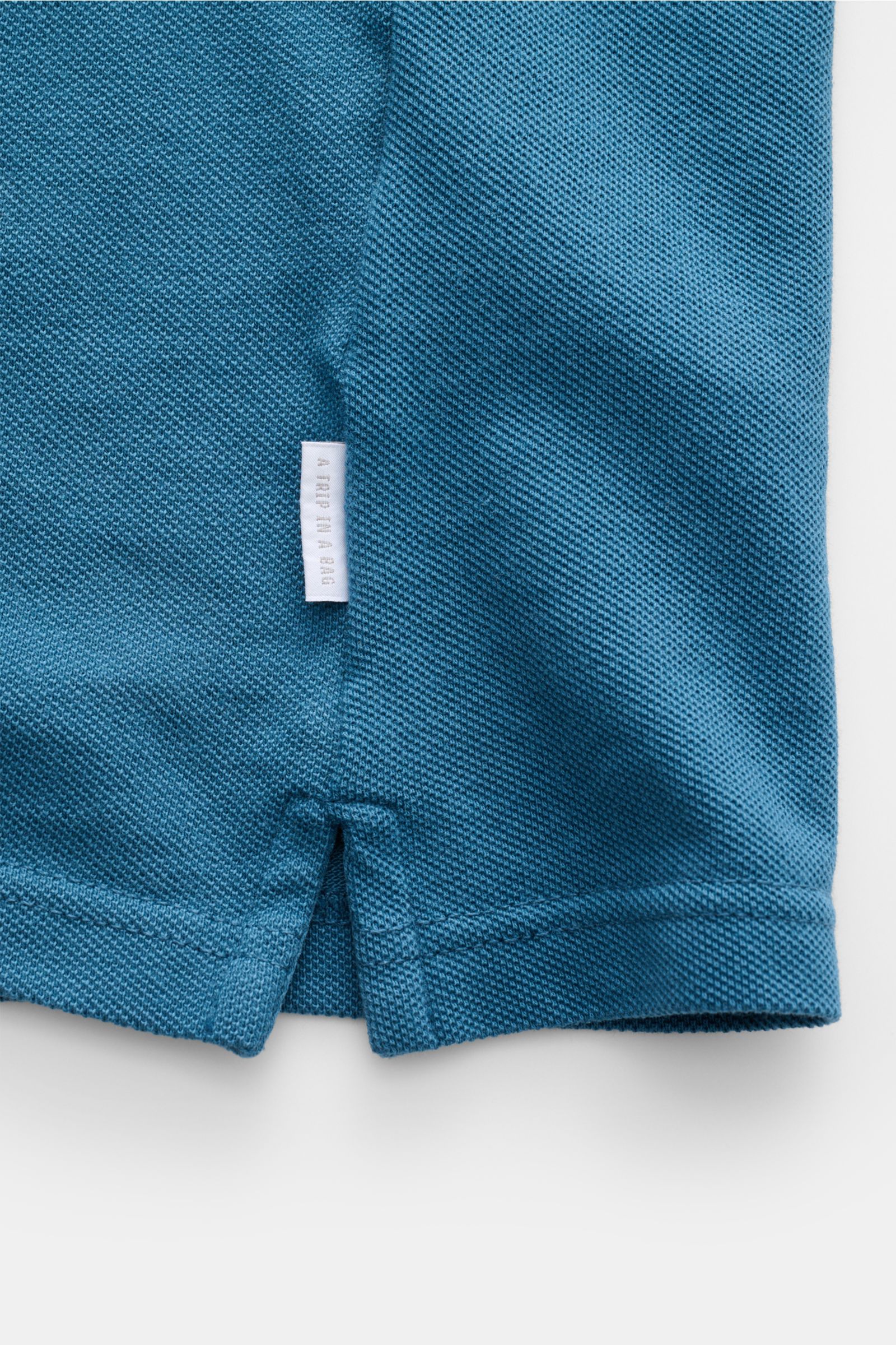 Nahaufnahme des 04651/ A trip in a bag Poloshirt 'Johnny' petrol, Piqué-Baumwolle, offene Kragenleiste, Weblabel seitlich, Saumschlitze.