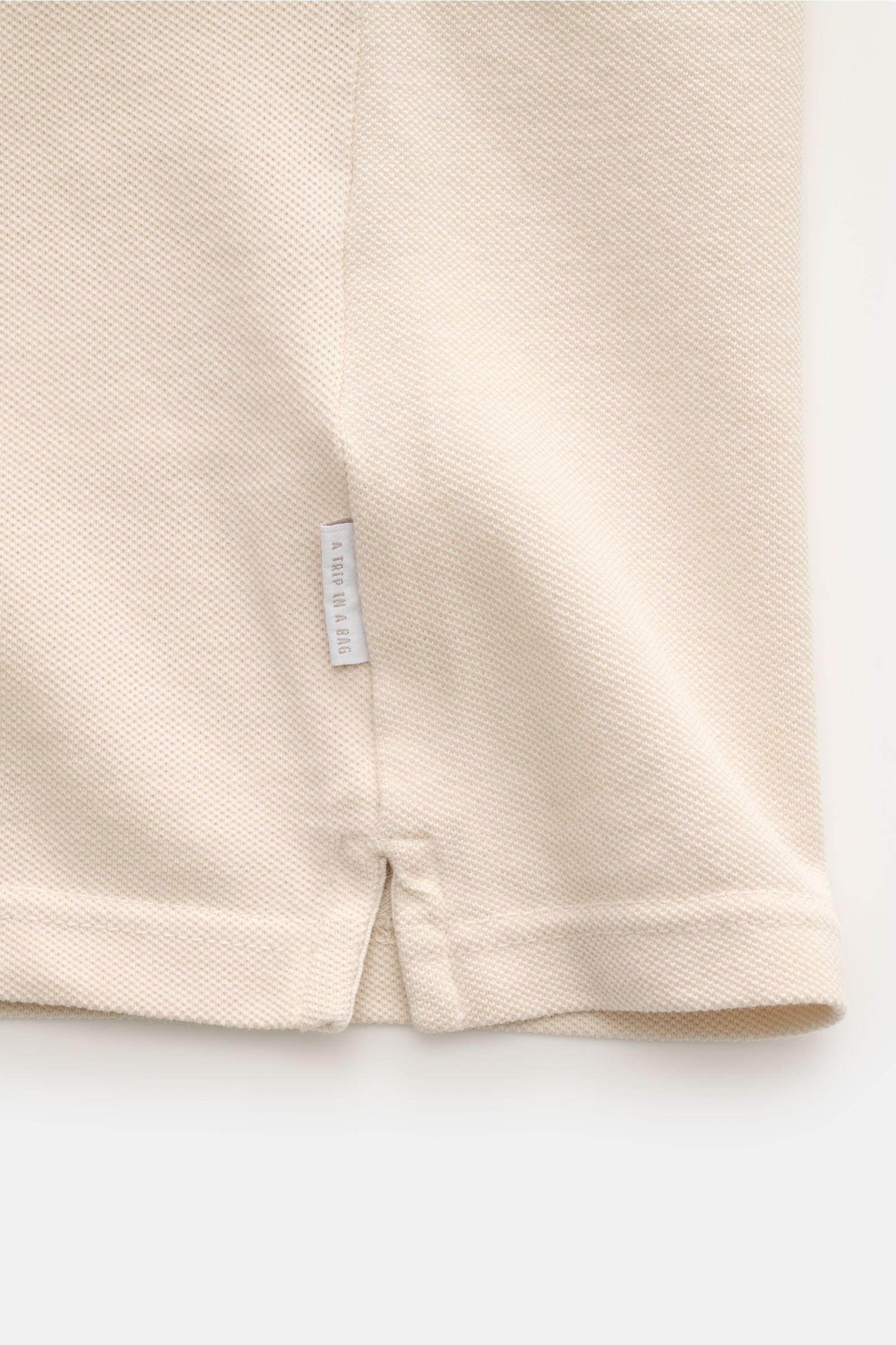 Close-up front view of the lower hem and side seam of the 04651/ A trip in a bag Poloshirt 'Johnny' sand, showing piqué cotton fabric with a small web label. Für lässige Freizeitlooks: 04651/ A TRIP IN A BAG bringt mit dem Poloshirt aus reiner Baumwoll