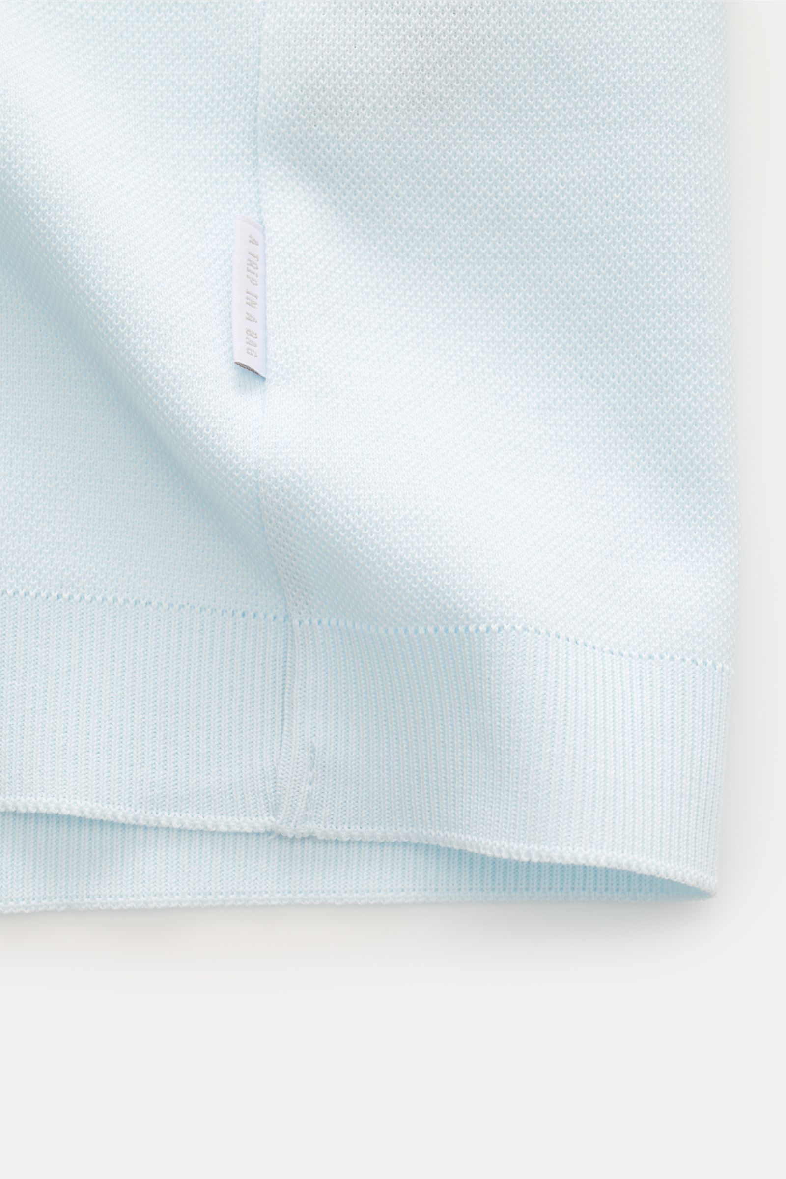Close-up from the side of the hem of the 04651/ A trip in a bag Poloshirt 'TP' pastellblau showing light blue cotton piqué fabric, ribbed cuffs, and a small white woven label.

Ob im Urlaub oder Office: 04651/ A TRIP IN A BAG bringt mit dem Poloshirt a