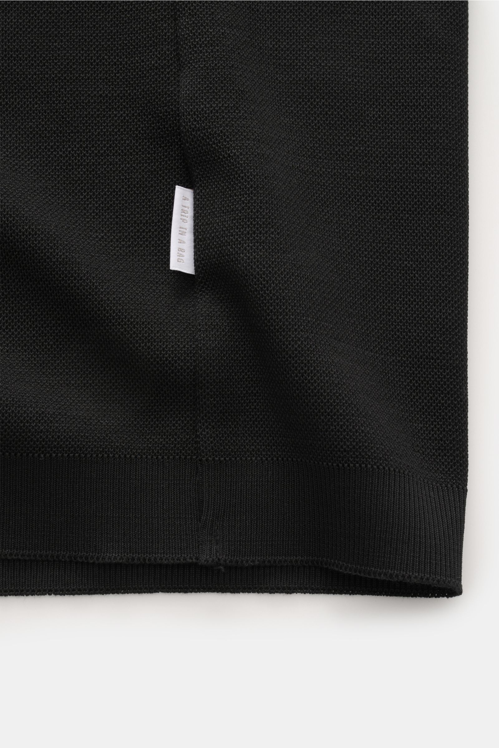 Close-up bottom hem detail of the 04651/ A trip in a bag Poloshirt 'TP' schwarz, showing black pure cotton piqué fabric, soft touch, regular fit, classic polo collar, smooth button placket, small side seam label, and comfortable ribbed cuffs, photographed