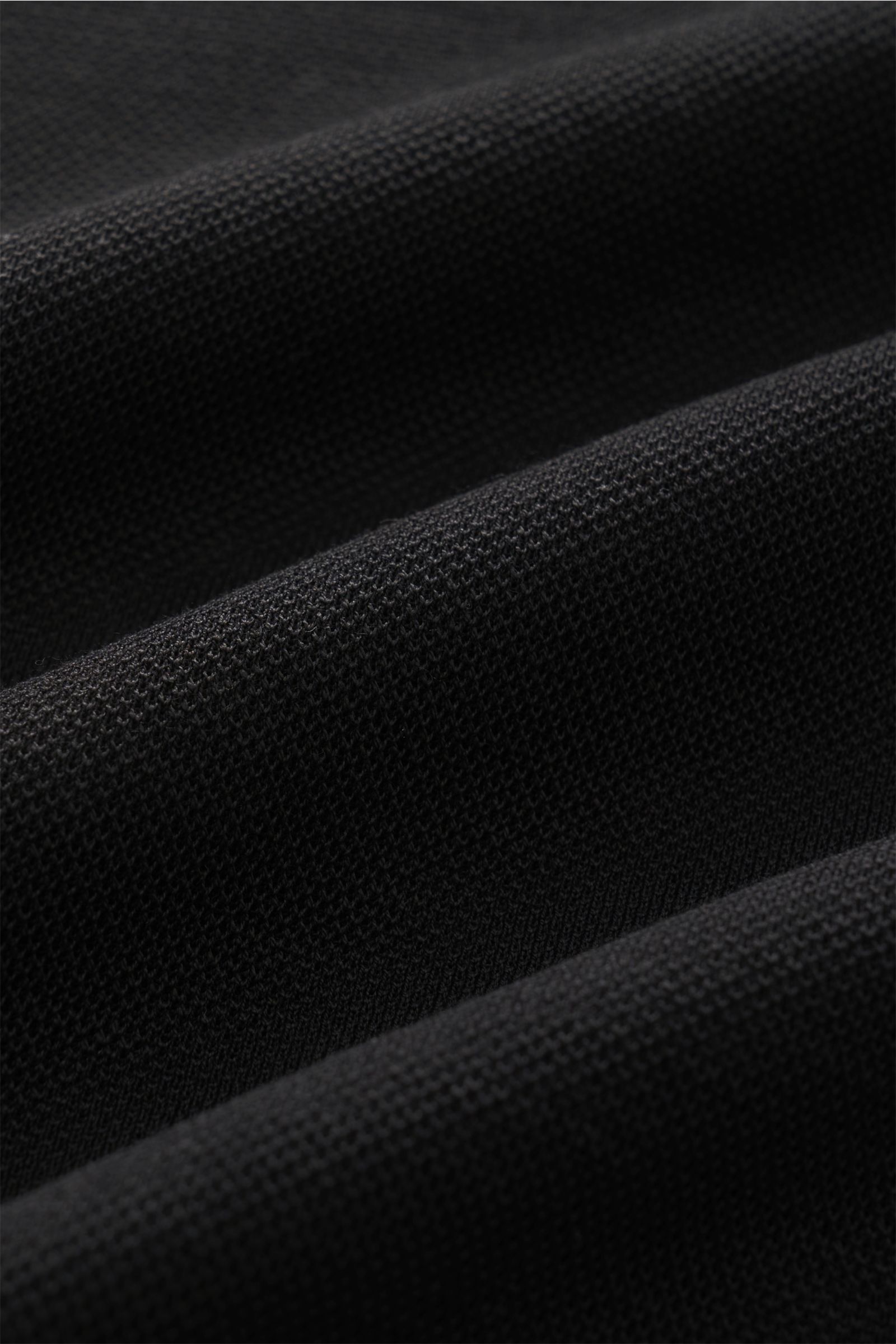 Close-up front perspective of black piqué cotton fabric showing texture and weave, from 04651/ A trip in a bag Poloshirt 'TP' schwarz. Ob im Urlaub oder Office: 04651/ A TRIP IN A BAG bringt mit dem Poloshirt aus reiner Baumwolle lässiges Sommergefühl 