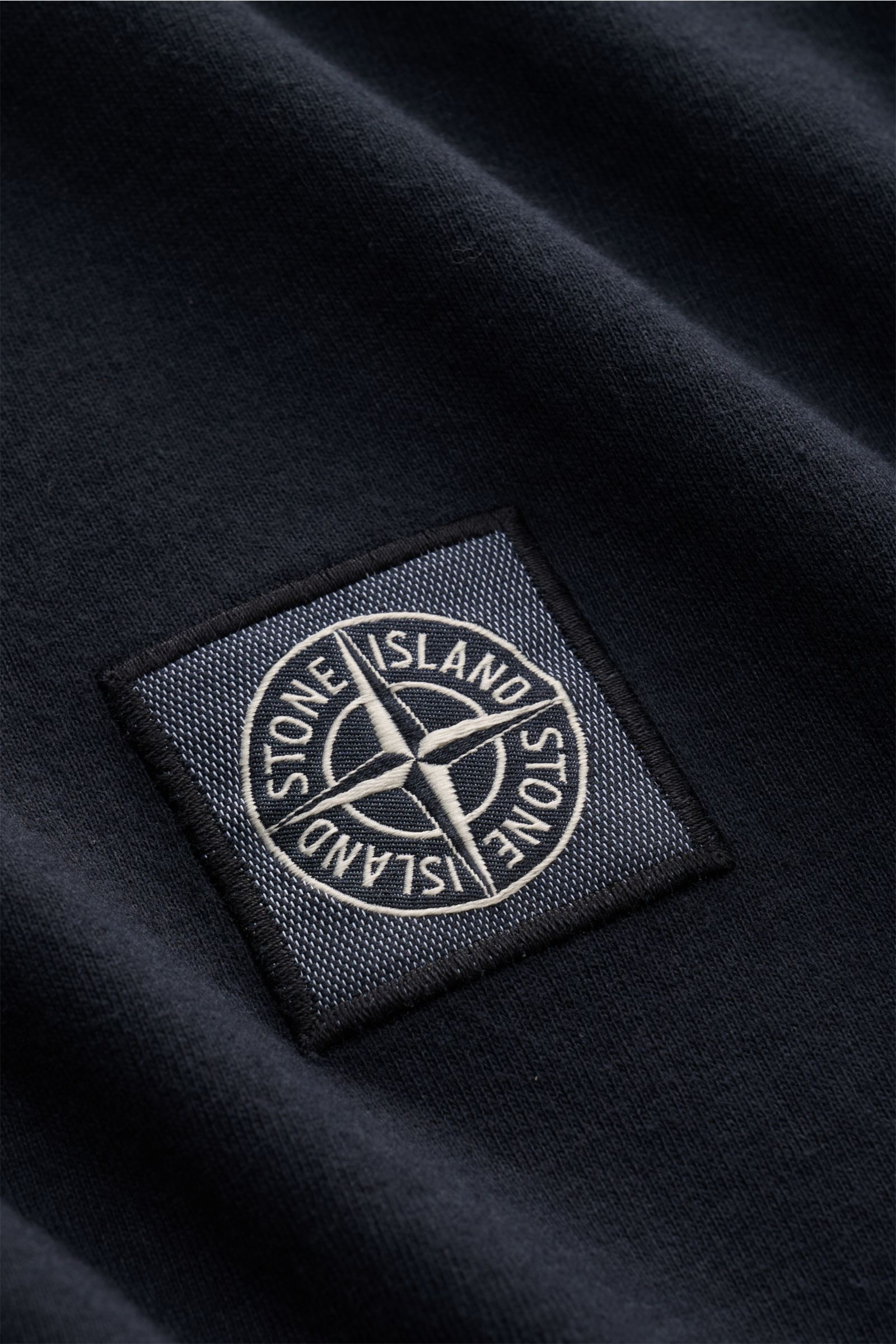 Nahaufnahme des Stone Island Rundhals-T-Shirts navy mit charakteristischem Kompass-Logo, weichem Bio-Baumwoll-Jersey, markanten Overlock-Nähten und STONE ISLAND-Weblabel, fotografiert aus schräger Perspektive.