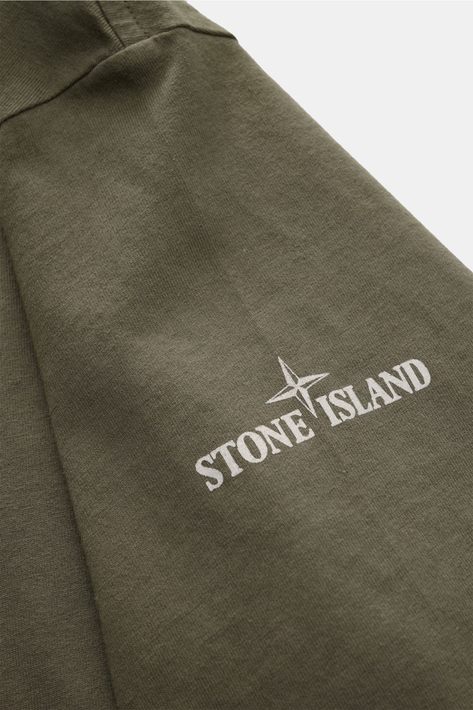 Close-up detail of the left sleeve of a Stone Island Rundhals-T-Shirt oliv, showing the white 'Stone Island' print on olive green jersey fabric. Jersey aus reiner Baumwolle, schwere Qualität, leicht griffige Haptik, garment dyed, Regular Fit, Rundhalsauss