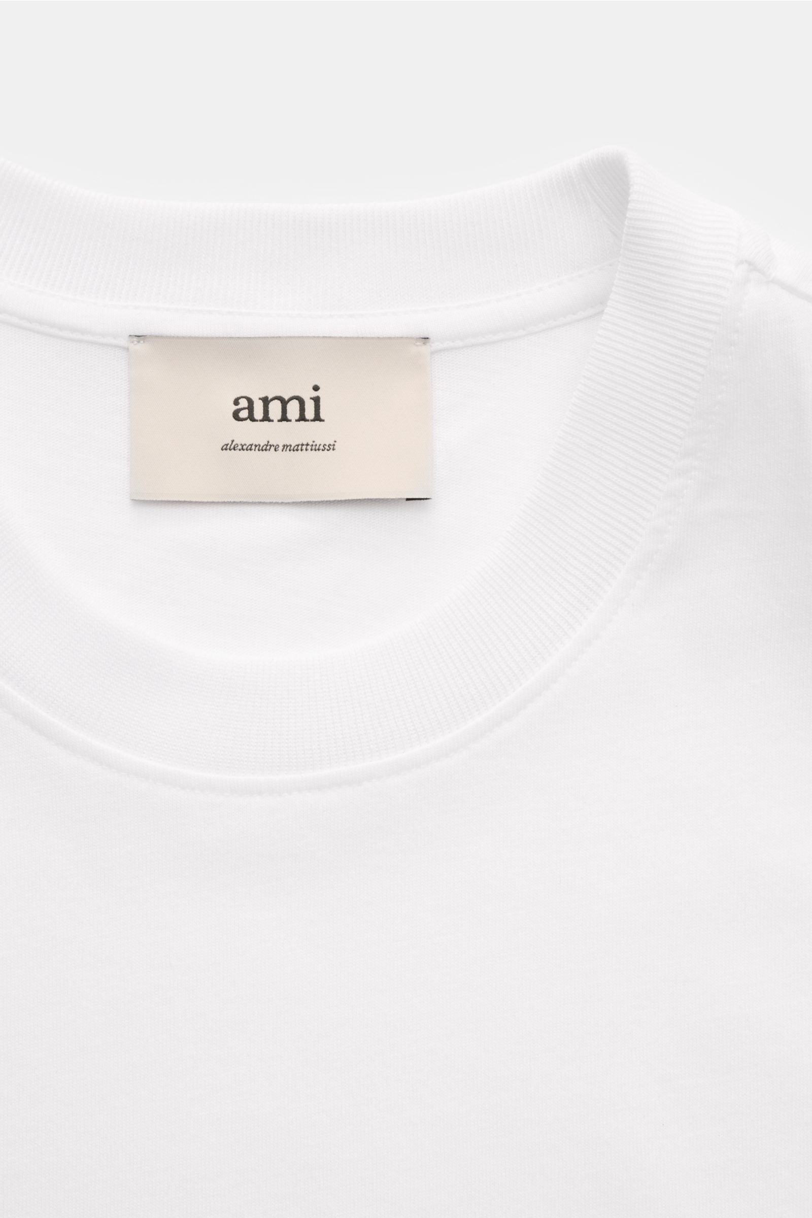 A close-up front view of the Ami Paris Rundhals-T-Shirt weiß showing the white round neckline and the beige label with 'ami alexandre mattiussi'. Klassisches Jersey-Basic vom französischen Label AMI PARIS: Dieses T-Shirt mit Rundhalsausschnitt ist aus 