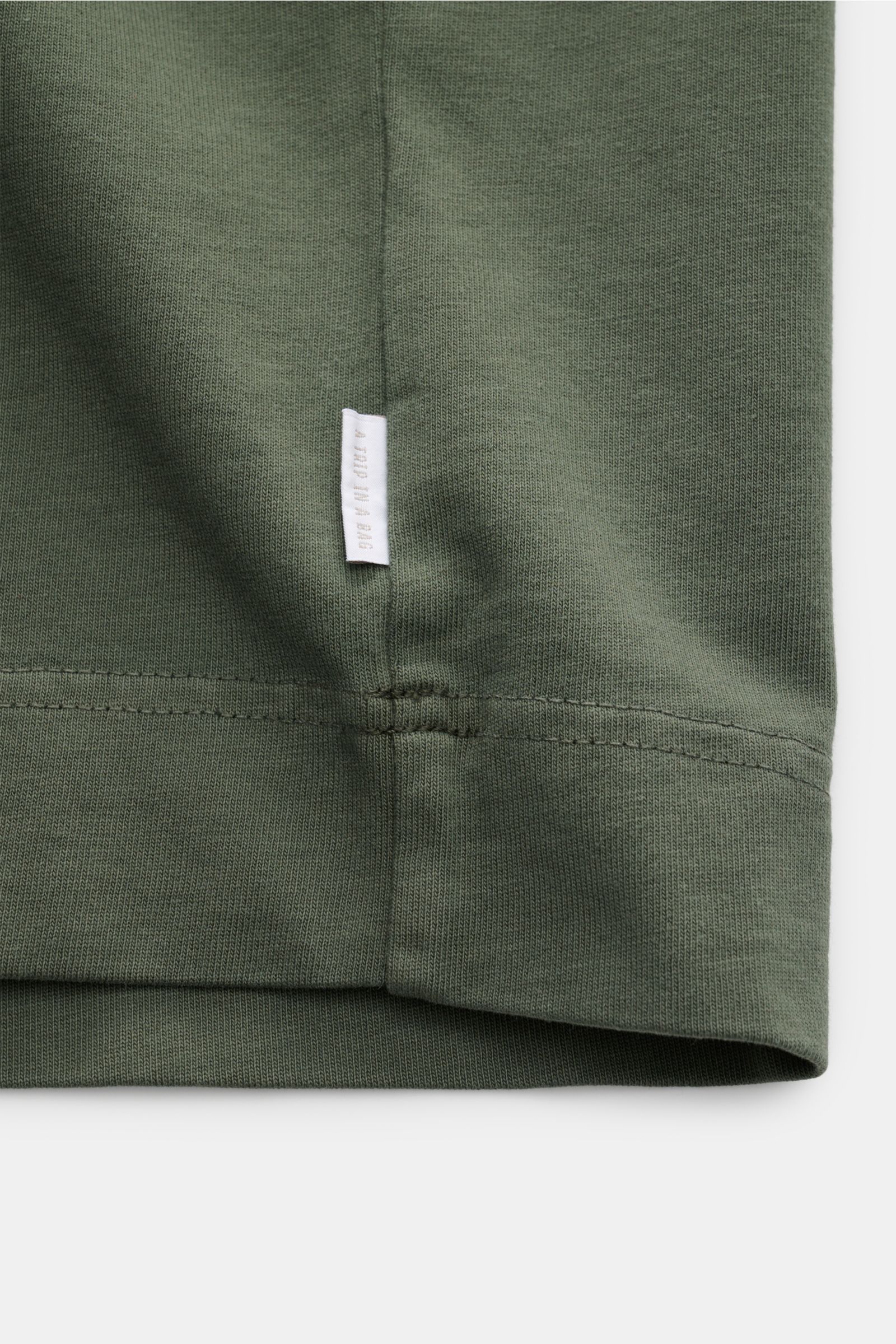 Close-up view of the hem and side seam of the 04651/ A trip in a bag Rundhals-T-Shirt 'Nunu Tee' graugrün in soft gray-green cotton jersey, showing the white web label in the side seam. Tauchen Sie ein in den relaxten Lifestyle von 04651/ A TRIP IN A B