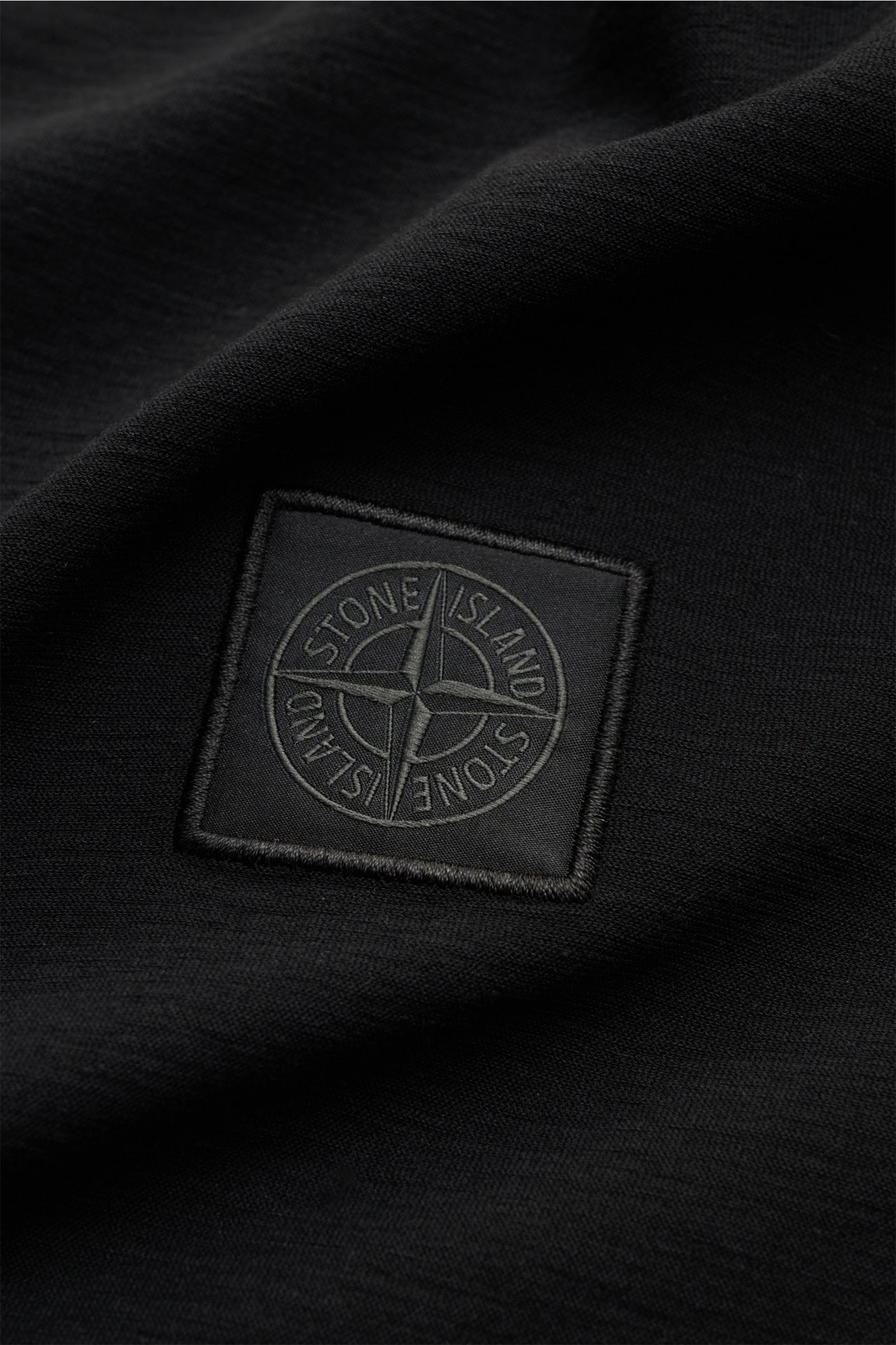 Close-up, top-down view of the Stone Island Rundhals-T-Shirt 'Ghost Piece' schwarz showing the tonal black STONE ISLAND-Weblabel on soft, slightly textured black cotton jersey fabric. STONE ISLAND bietet mit dem Rundhals-T-Shirt aus elastischem Baumwol