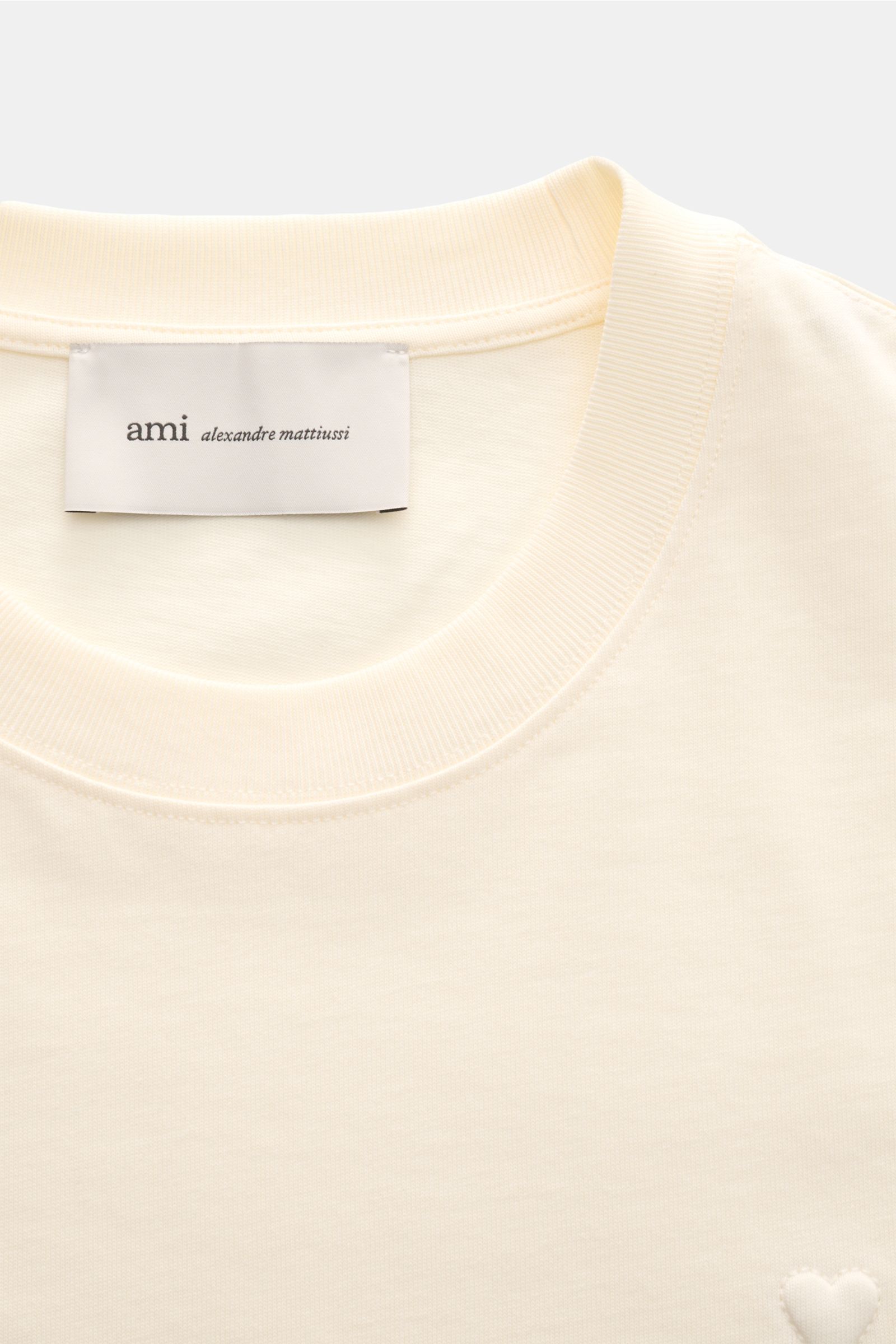Ami Paris Rundhals-T-Shirt creme, Nahaufnahme von Rundhalsausschnitt, Jersey aus Baumwolle, abgesteppte Herz-Logo und Label innen.
